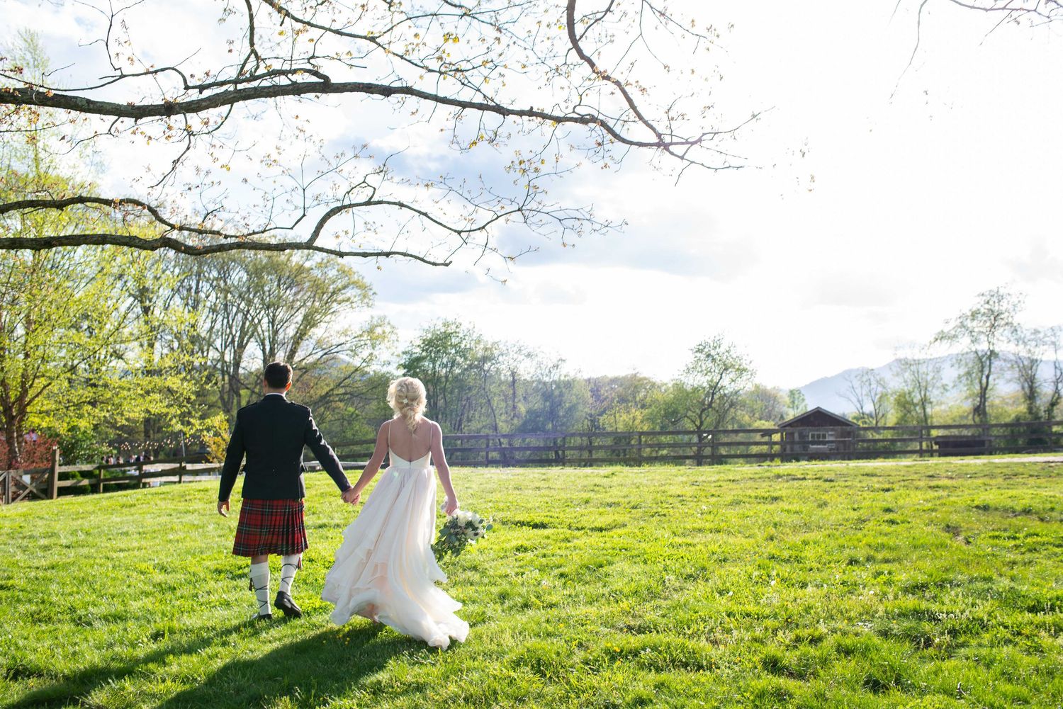 Bride in white gown and groom in Scottish kilt walk hand-in-hand across lush green field at outdoor wedding venue.