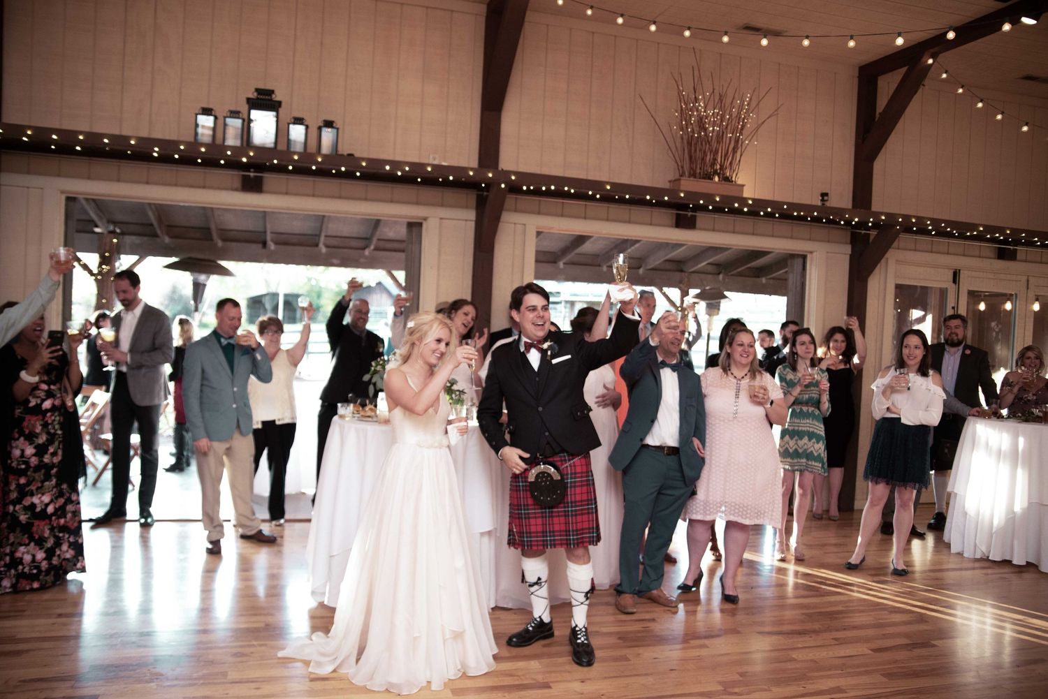 Bride in white gown and groom in red kilt celebrate with guests on wooden dance floor at rustic barn wedding reception.