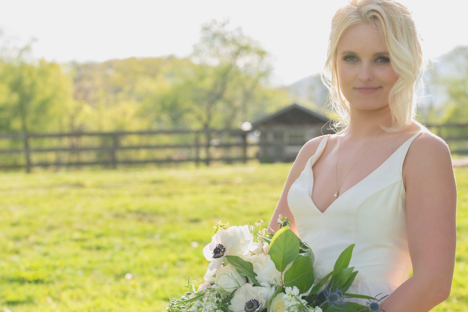 Blonde bride in white gown holding white floral bouquet with greenery, standing in sunny green field near rustic fence.