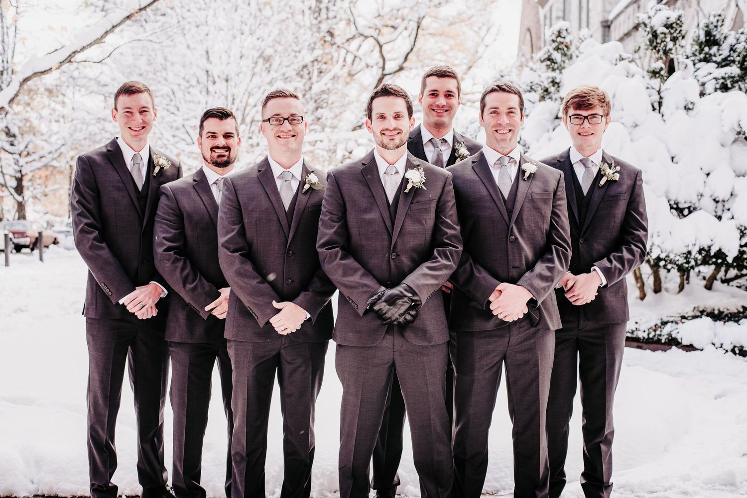 Seven groomsmen in matching dark suits pose outdoors in a snowy winter wedding photo.