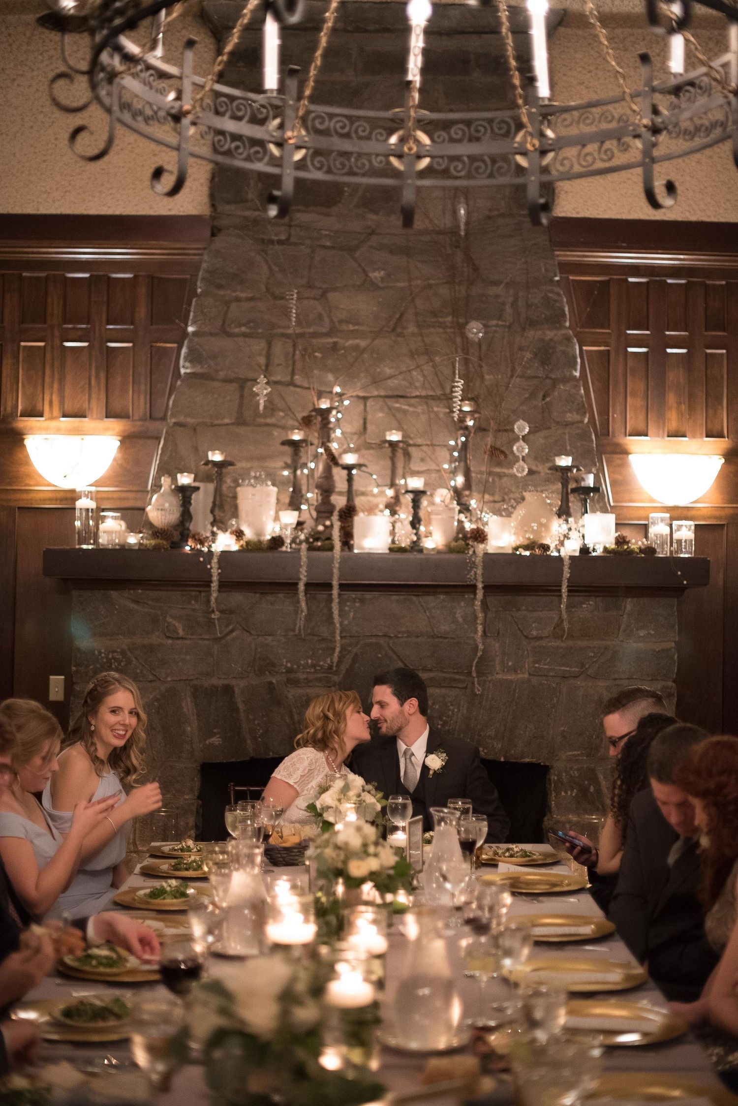 Romantic wedding reception dinner by candlelight with couple sharing a kiss near a stone fireplace mantel.