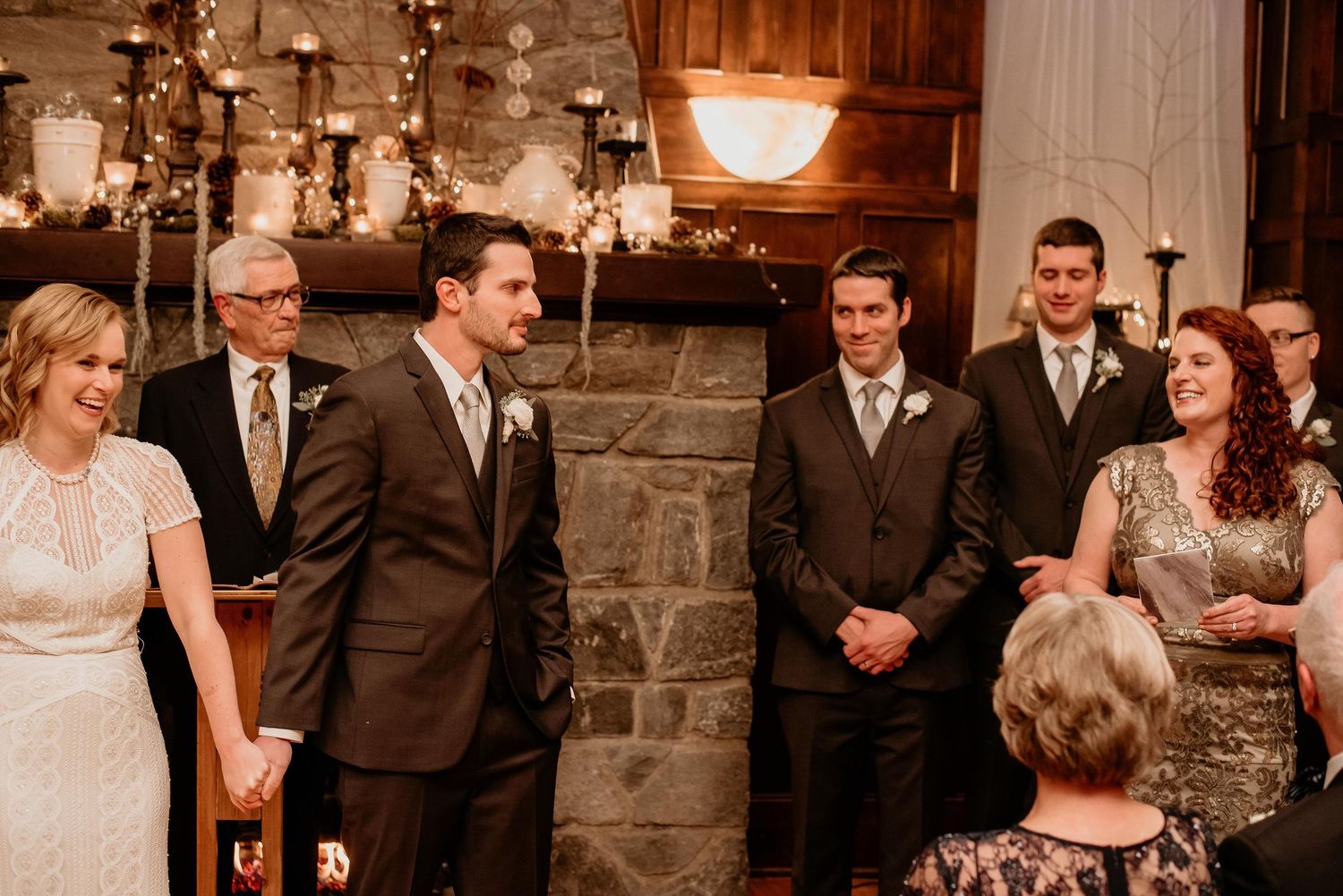 Wedding guests and groomsmen smiling during an indoor ceremony near a stone fireplace with candles.