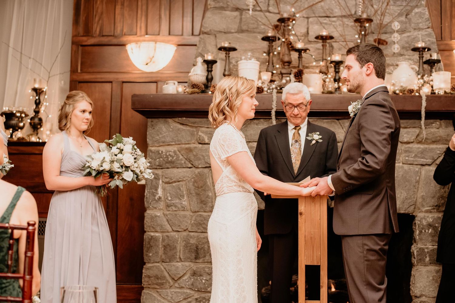 Bride and groom exchange vows at rustic stone fireplace altar with bridesmaid and officiant during indoor wedding ceremony.
