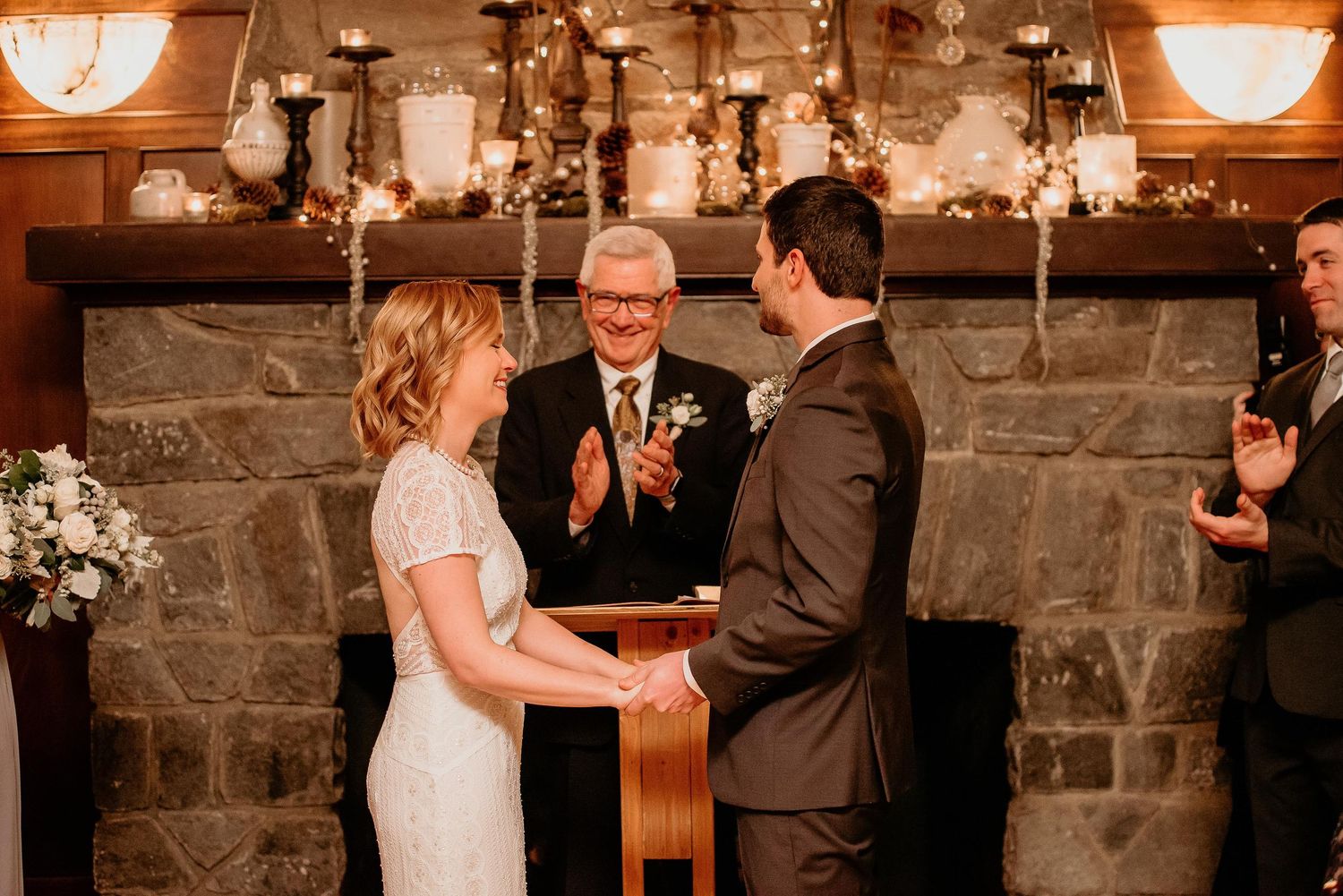 Couple exchanges vows at romantic indoor wedding ceremony by stone fireplace with officiant clapping.