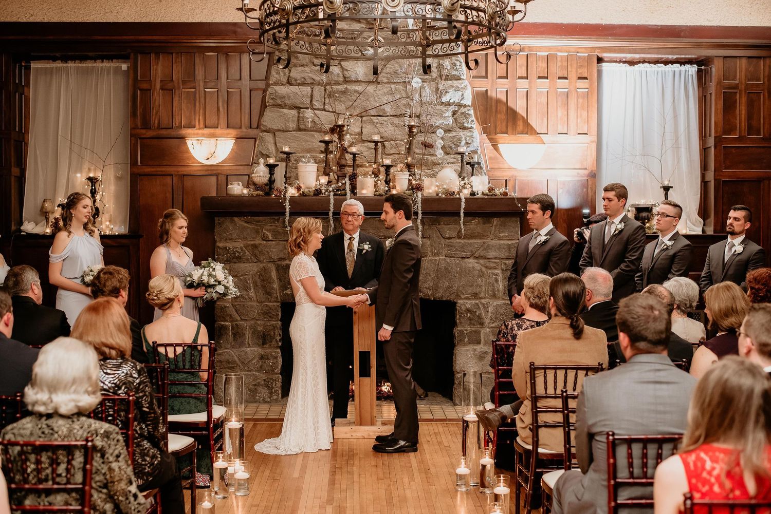 Elegant indoor wedding ceremony with couple exchanging vows before stone fireplace, chandelier, and wedding party in formal attire.