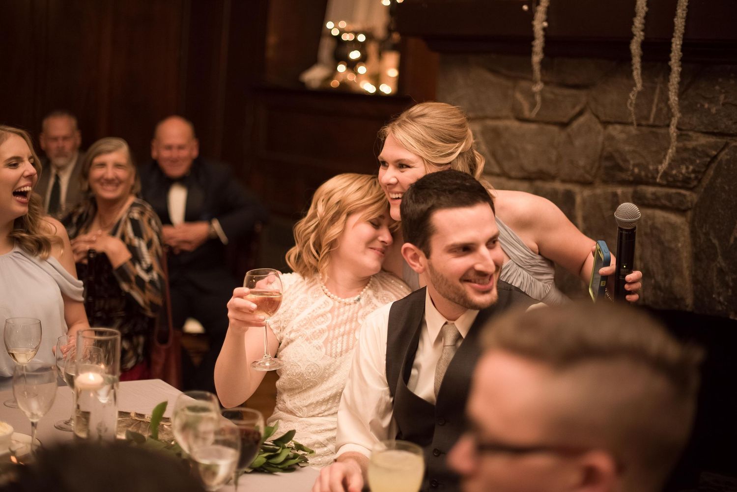 Wedding guests laughing and celebrating at a reception dinner inside a rustic venue with stone fireplace.