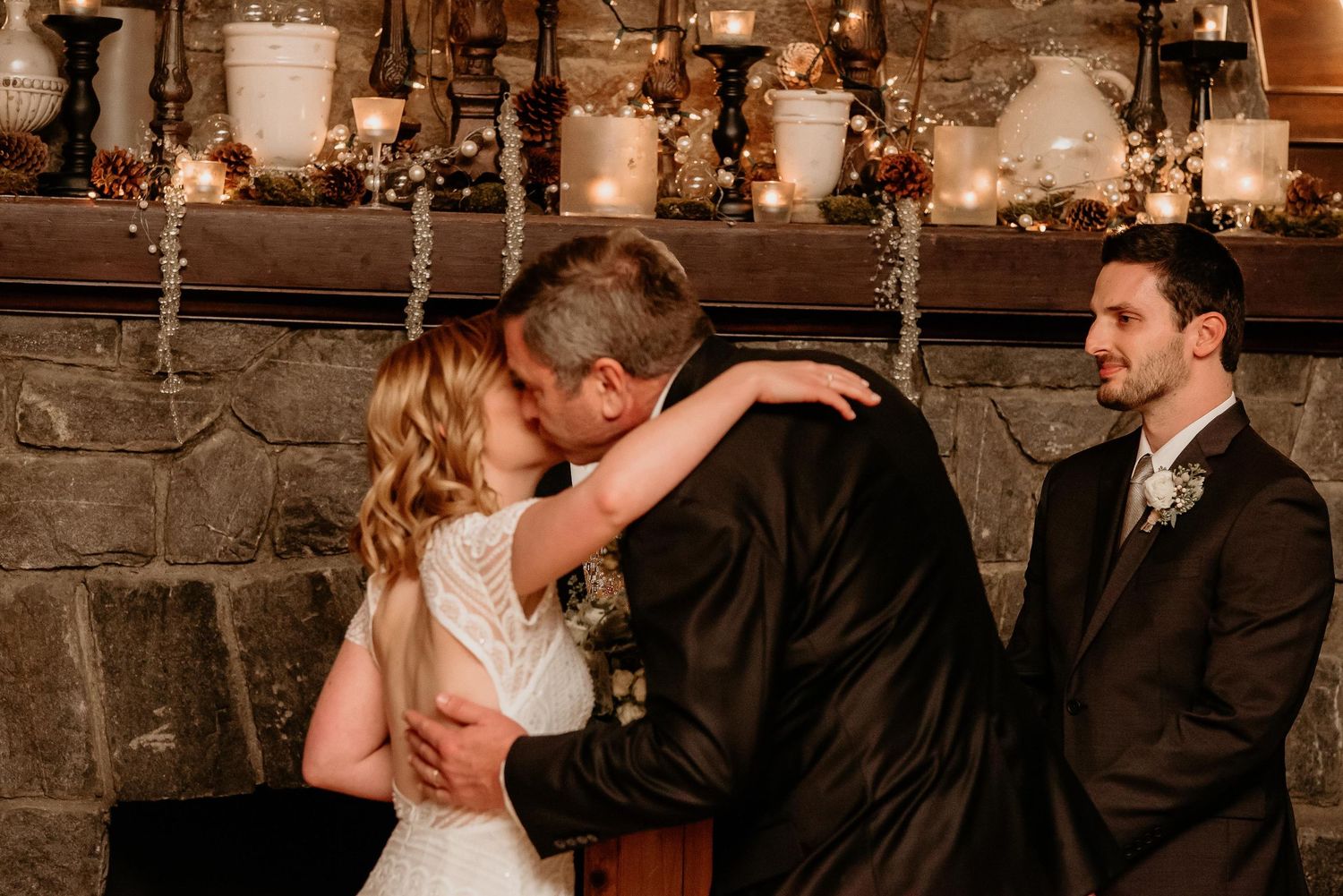 Bride and groom share first kiss at rustic stone fireplace wedding ceremony with candles glowing behind them.