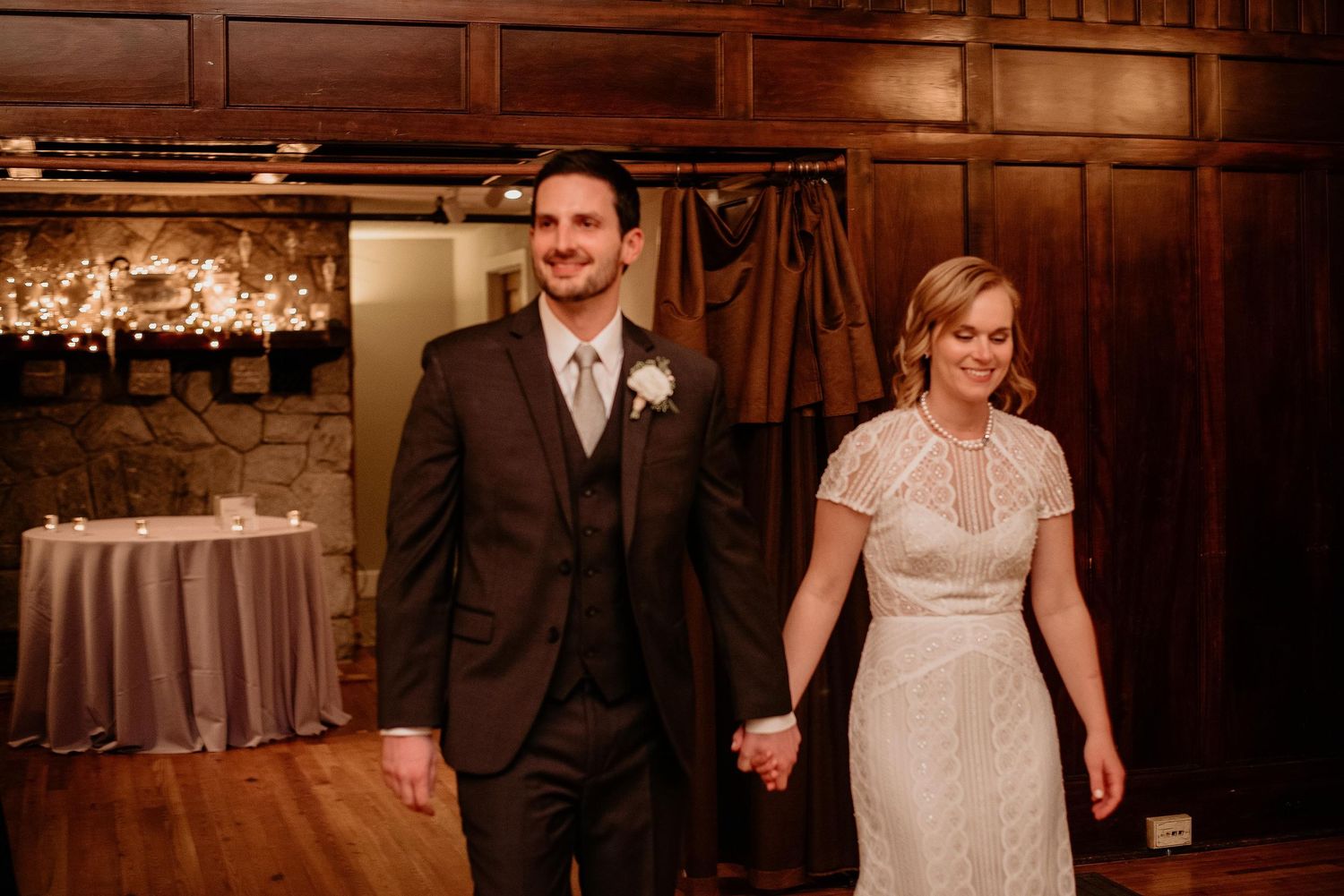 Bride and groom holding hands smiling as they enter their wedding reception in a warmly lit wood-paneled venue.