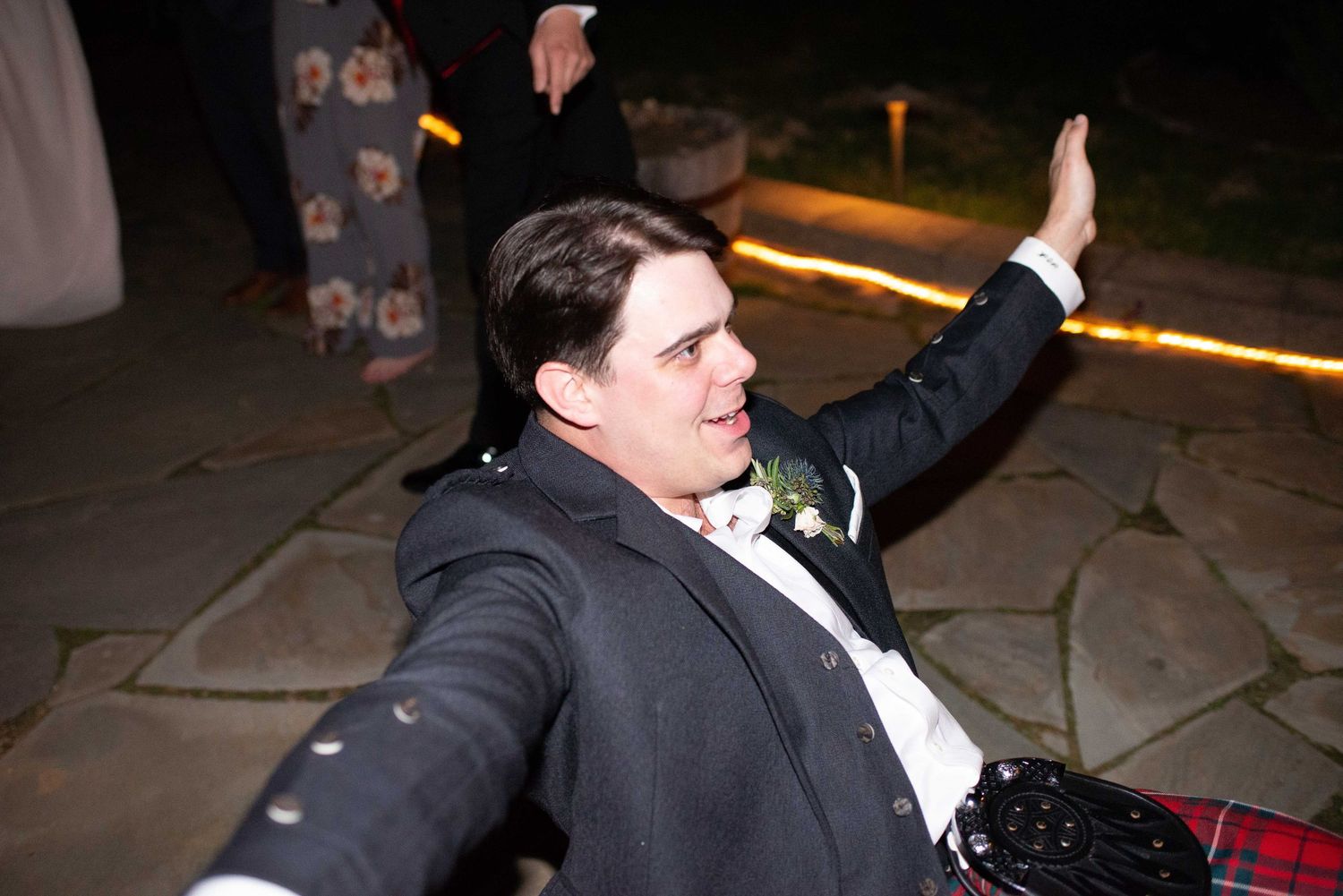 Man in suit with boutonniere sitting on ground at night, raising hand and smiling at outdoor wedding reception.