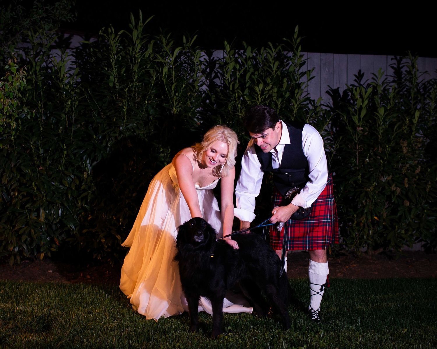 Bride in gold gown and groom in red tartan kilt petting a black dog outdoors at night.