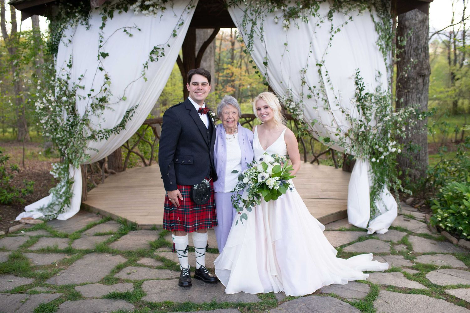 Wedding couple in Scottish kilt and bridal gown with elderly guest under decorated garden gazebo.