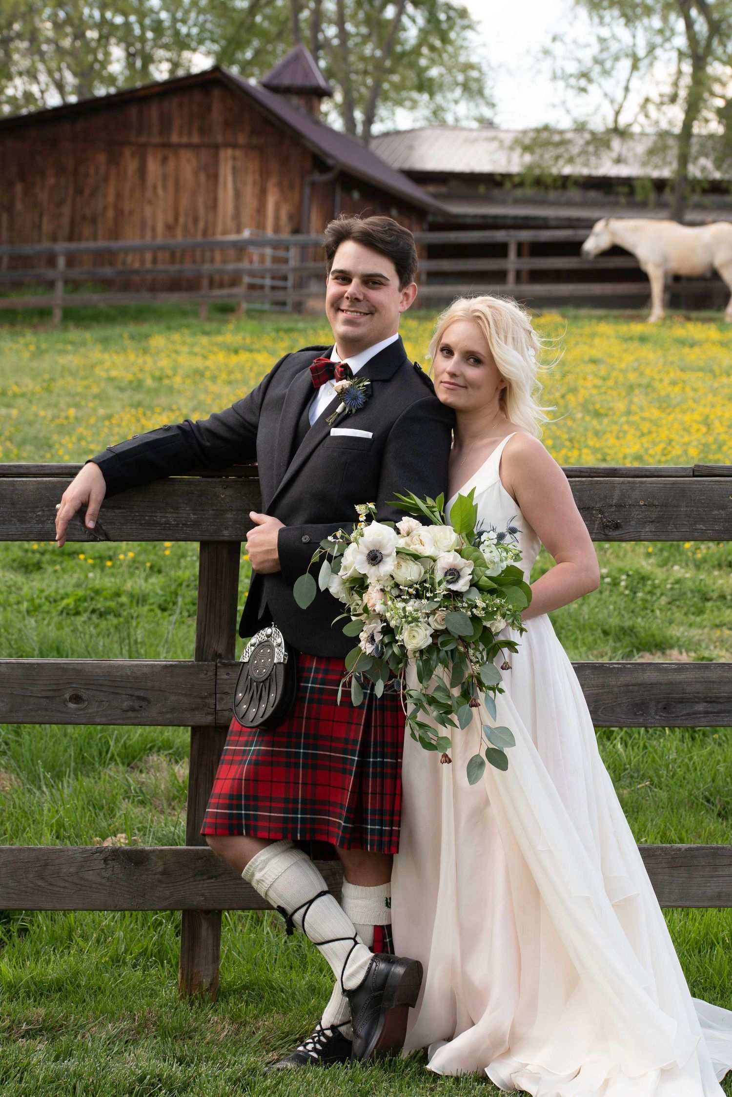 Bride and groom in Scottish kilt pose by rustic fence at farm wedding with white bouquet and horse in background.