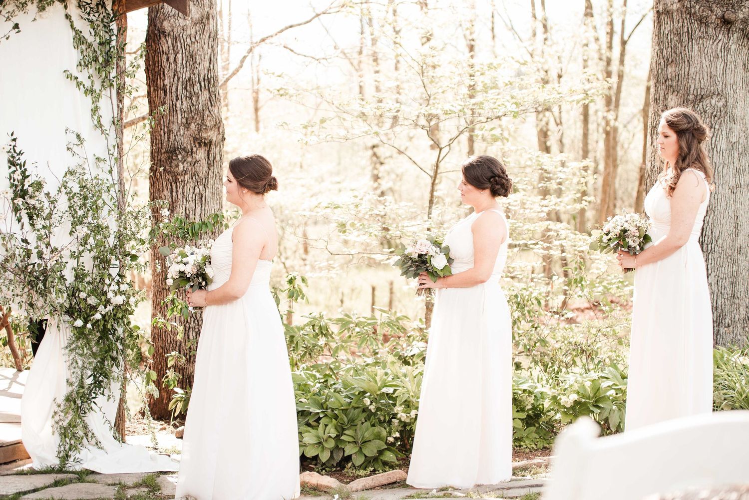 Three bridesmaids in white gowns holding floral bouquets stand outdoors among lush greenery at a woodland wedding.