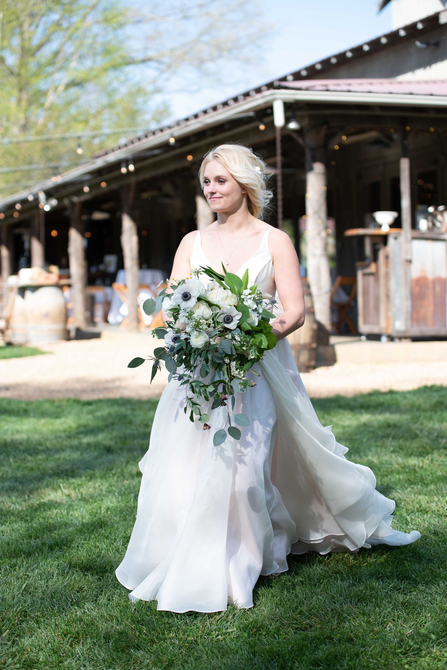 Bride in white gown holding lush white and green bouquet walking on grass at rustic barn wedding venue.