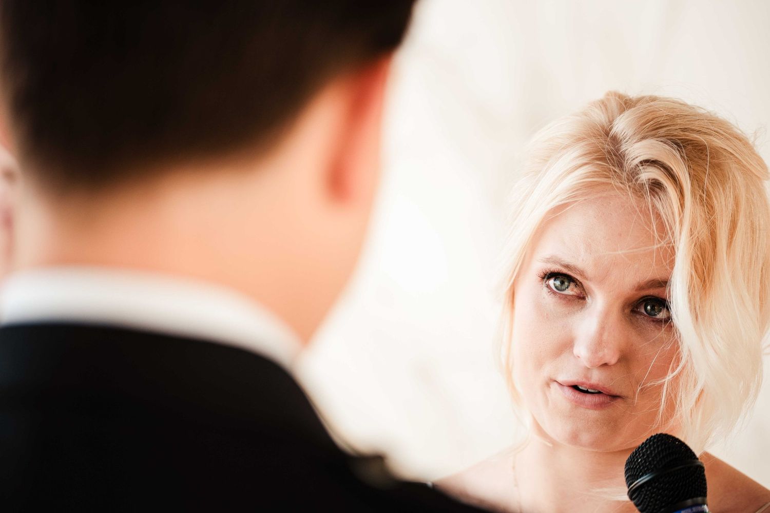 Blonde woman with blue eyes speaking into a microphone at a formal event, facing a man in a suit.