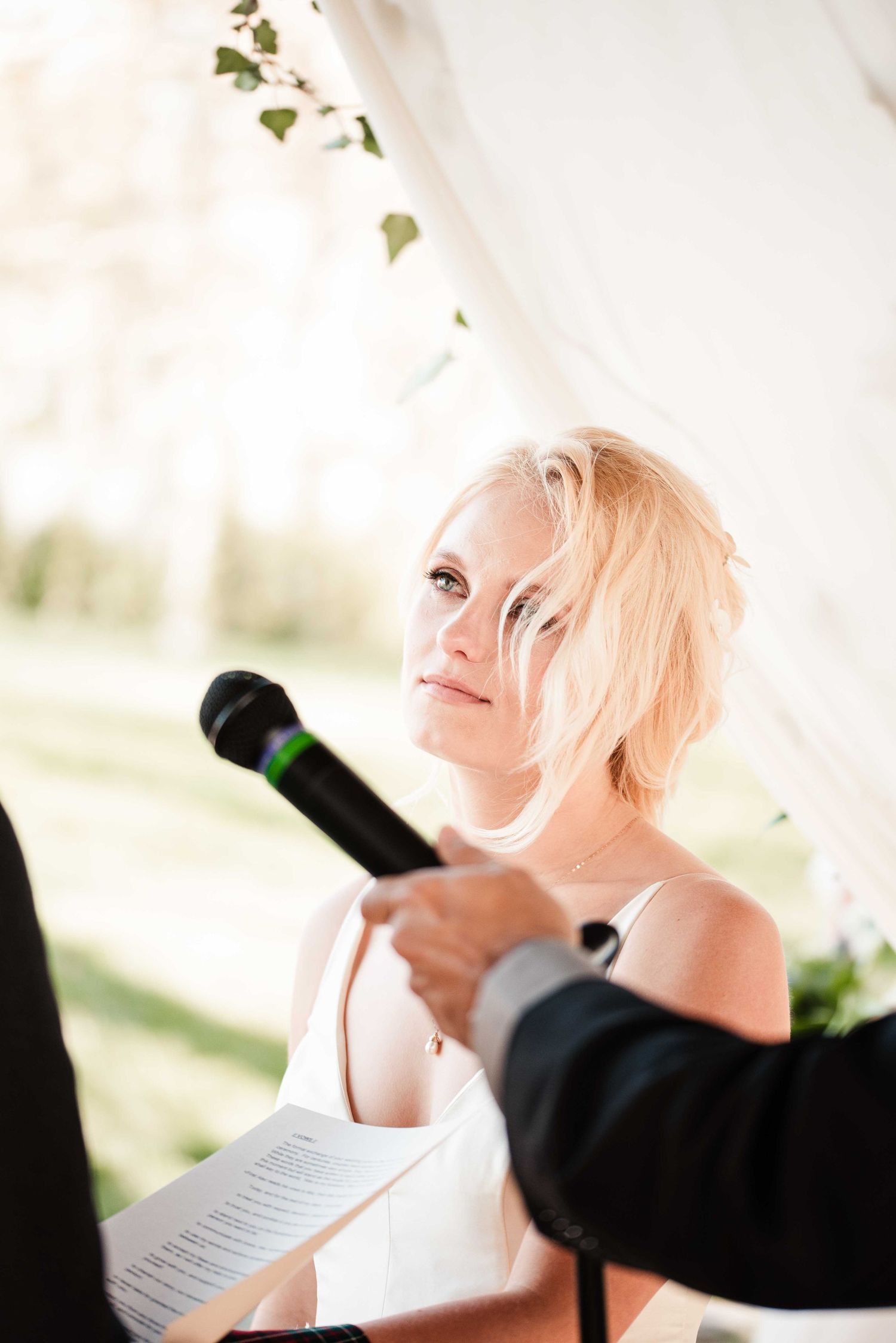 Blonde bride holding papers listens attentively during outdoor wedding ceremony with microphone nearby.