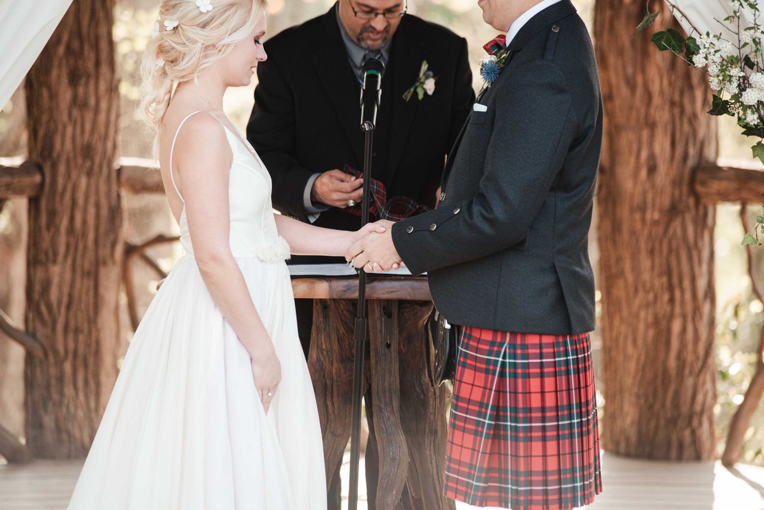 Bride and groom exchange vows outdoors, groom wearing red tartan kilt, officiant standing behind wooden altar.