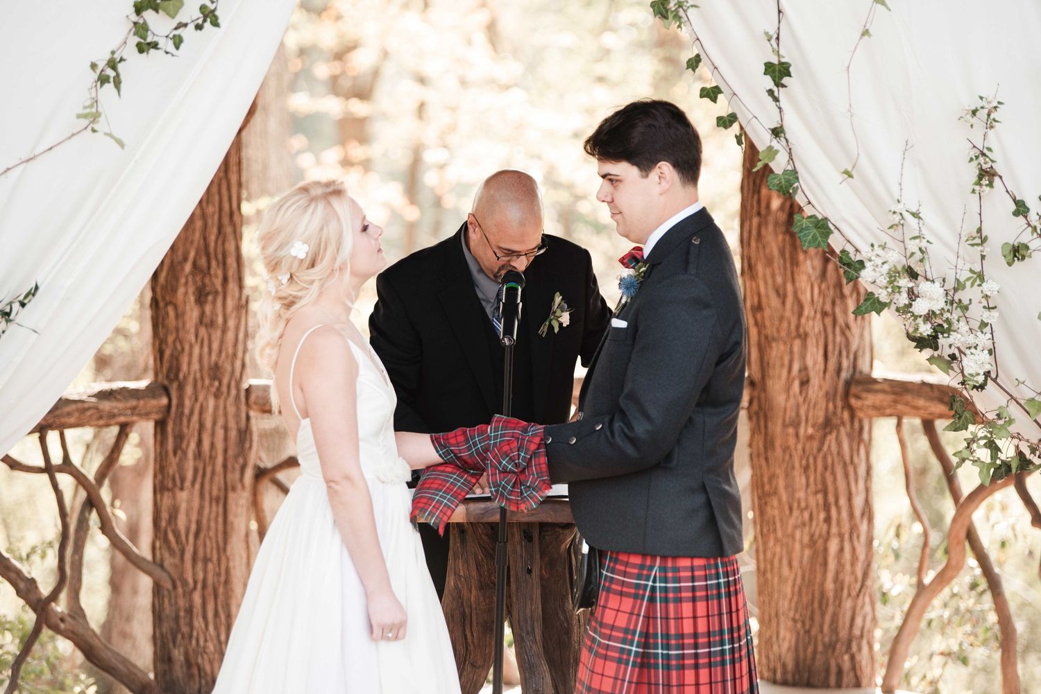 Groom in red tartan kilt exchanges vows with bride at rustic outdoor wedding ceremony with wooden arch.