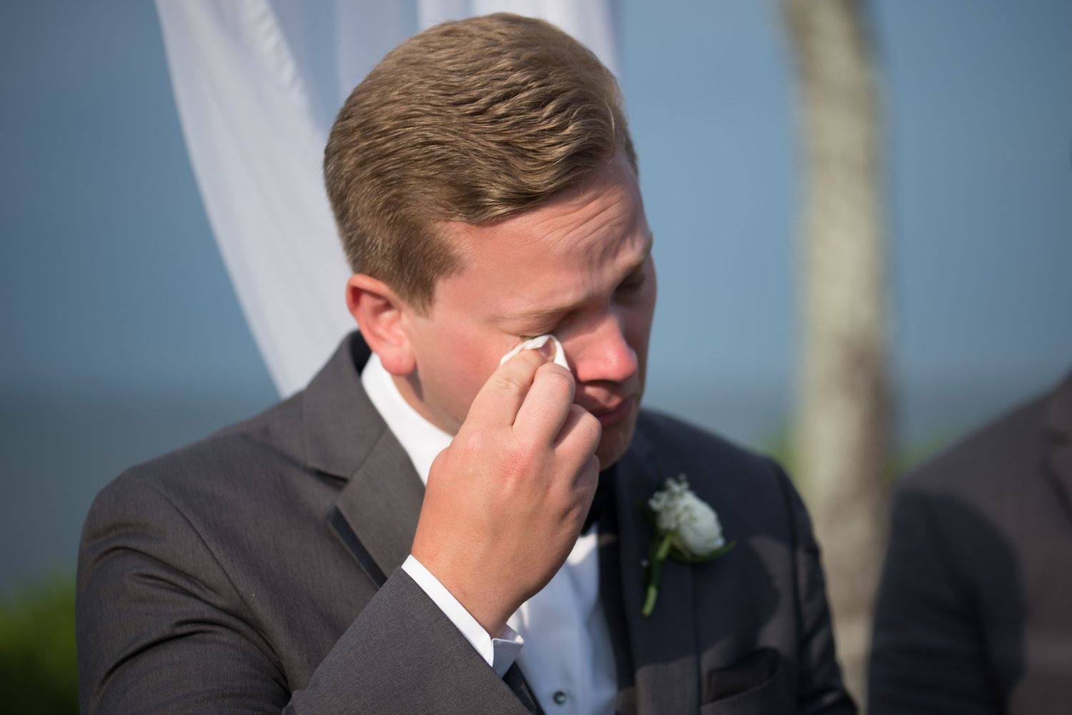 Emotional groom wiping tears during outdoor beach wedding ceremony.