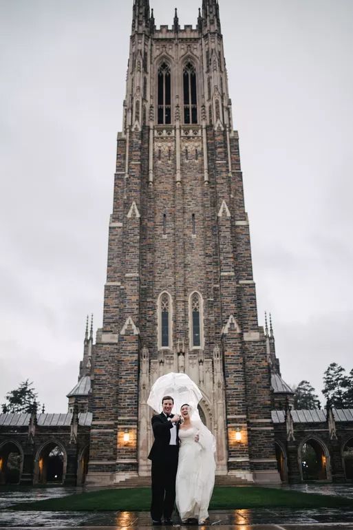 Bride and groom stand under umbrella before Gothic stone cathedral tower on rainy wedding day.