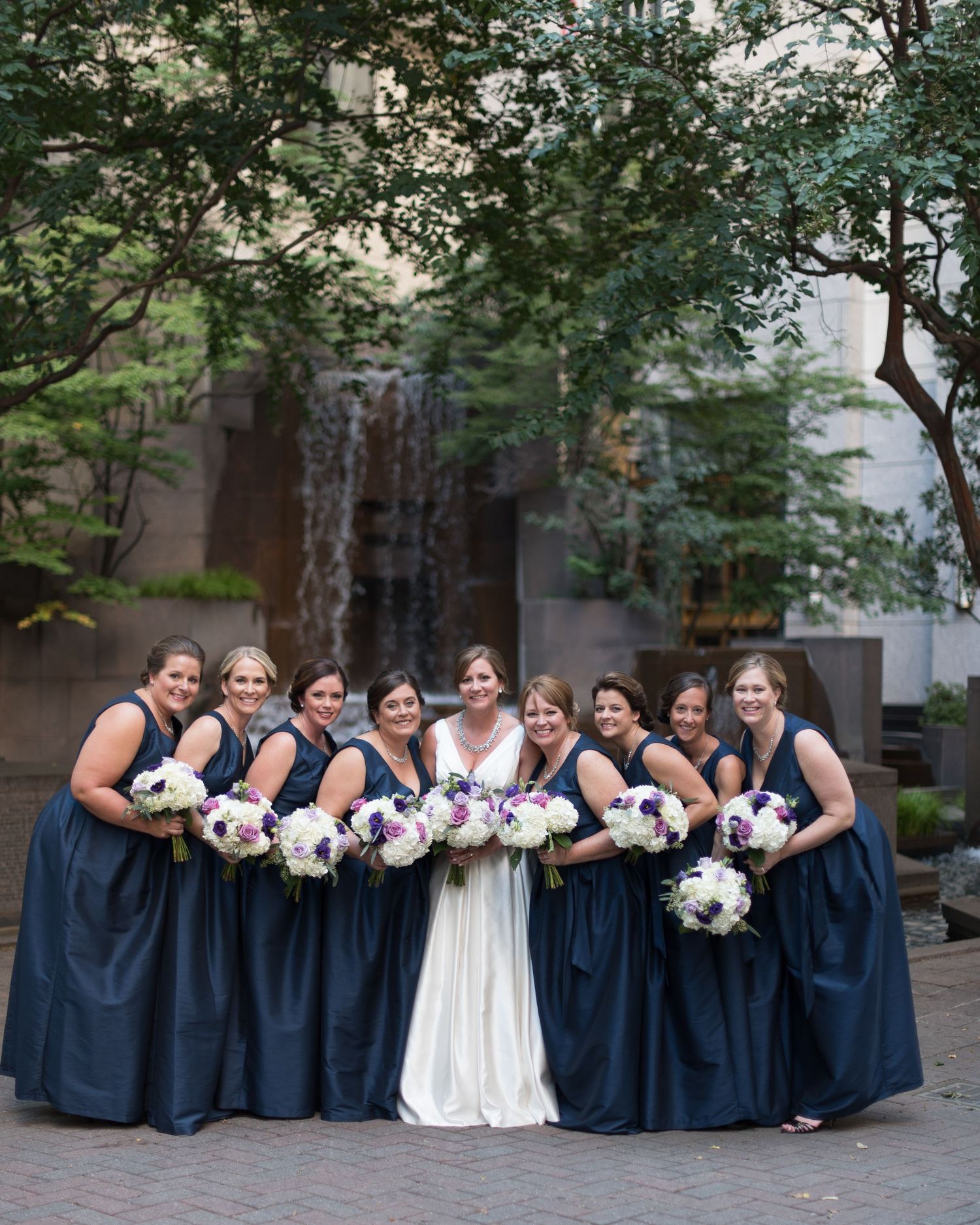 Bride with bridesmaids in navy dresses holding white bouquets outdoors.