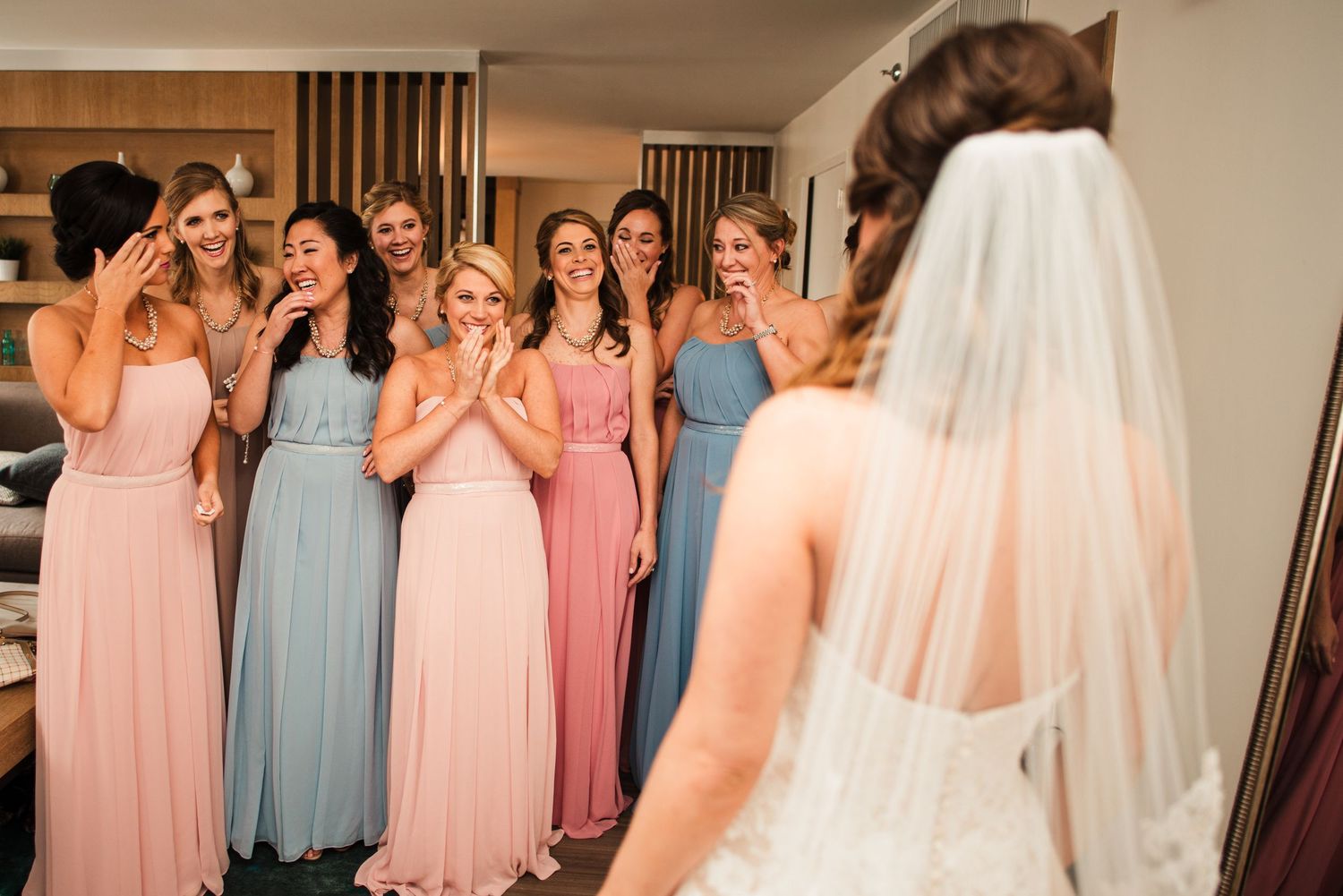 Bridesmaids in pink and blue gowns react with joy seeing the bride in her wedding dress for the first time.
