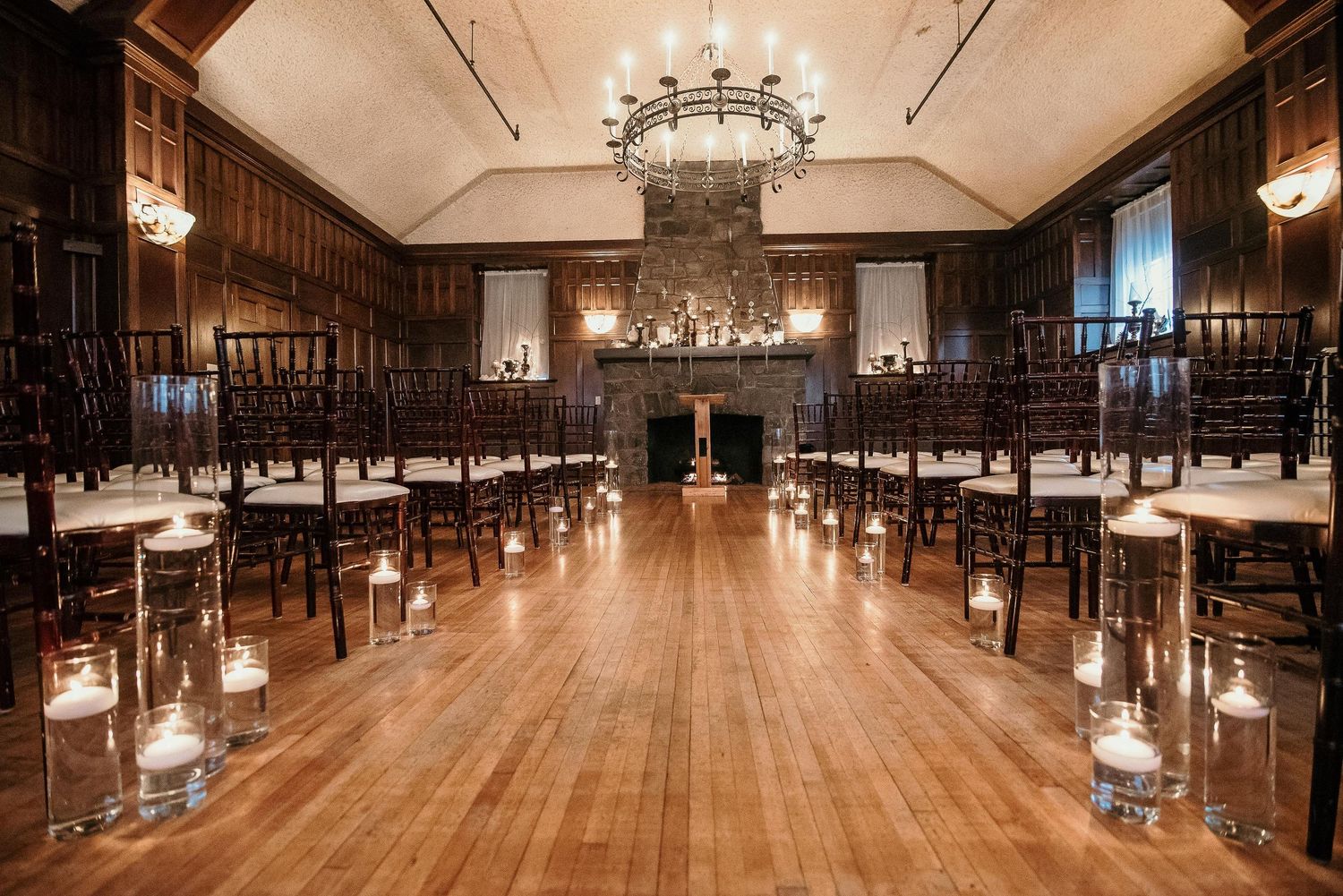 Elegant wedding ceremony venue with wood-paneled walls, candlelit aisle, chiavari chairs, and chandeliers on hardwood floors.