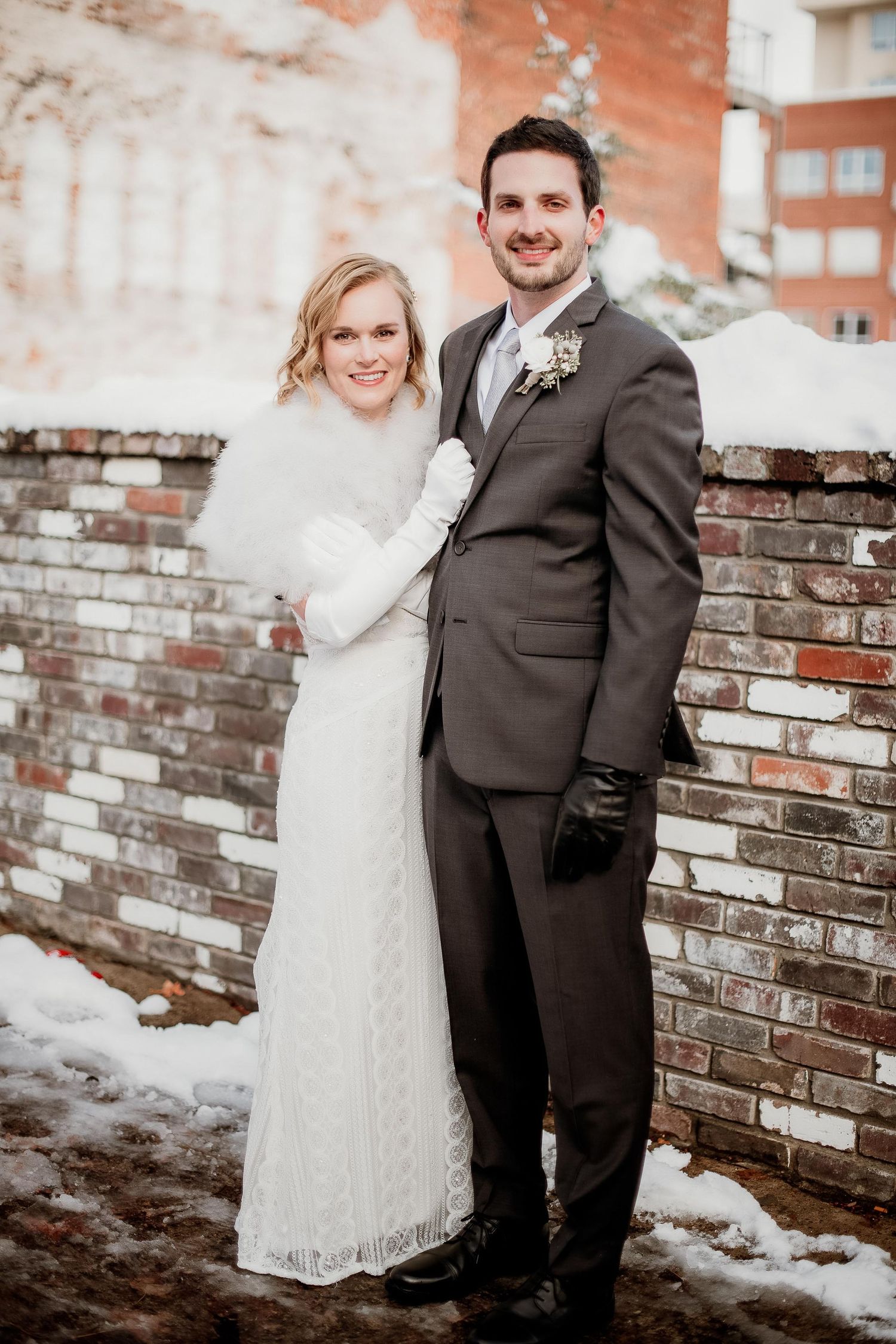 A bride and groom pose together in winter attire beside a brick wall with snow on the ground.