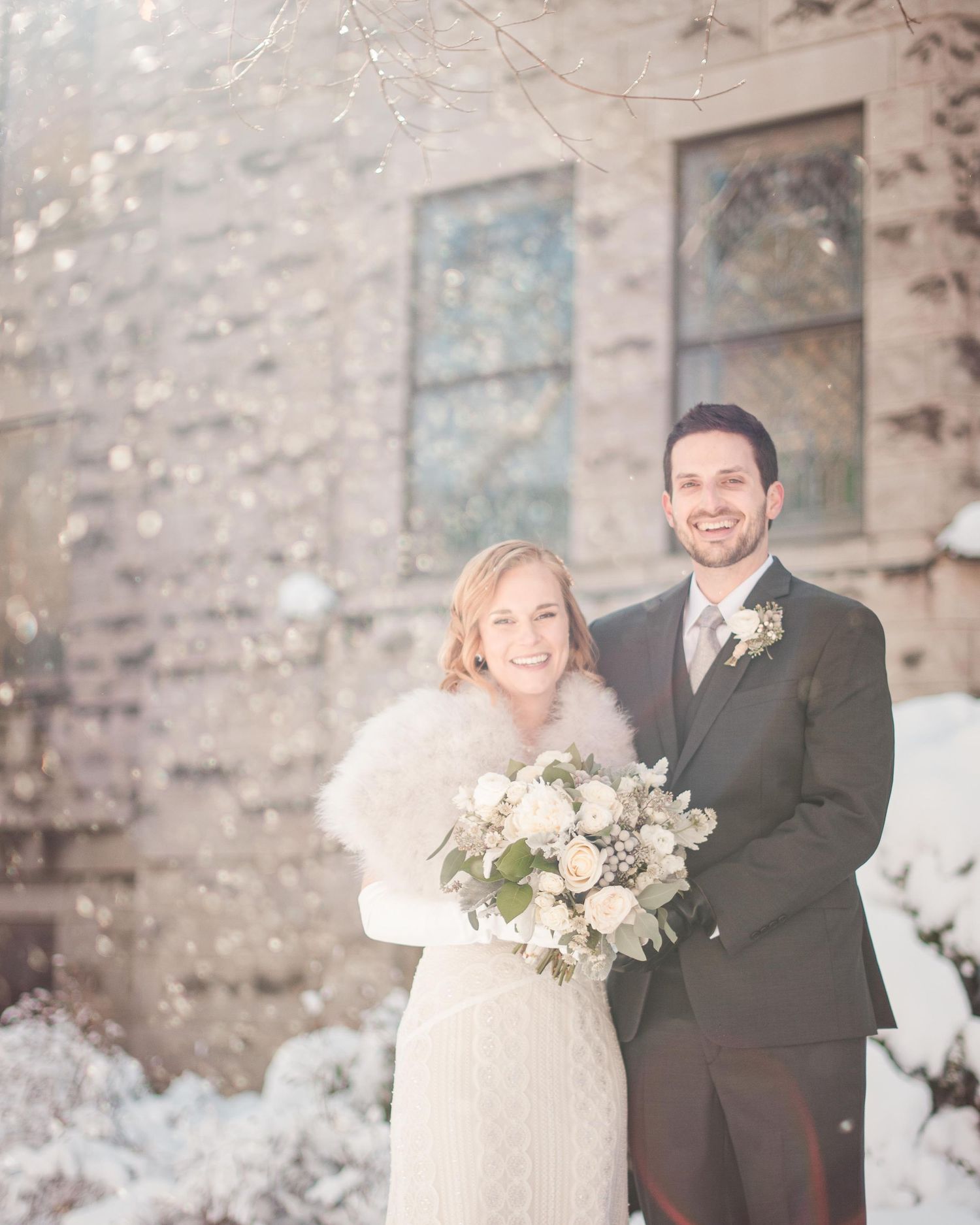 Happy couple posing outdoors in snow on winter wedding day, bride in white gown with fur wrap holding bouquet.