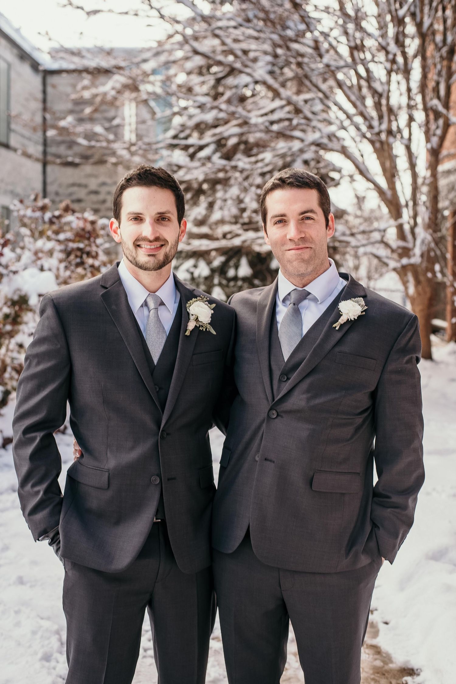 Two groomsmen in dark suits with boutonnieres pose outdoors in a snowy winter wedding setting.