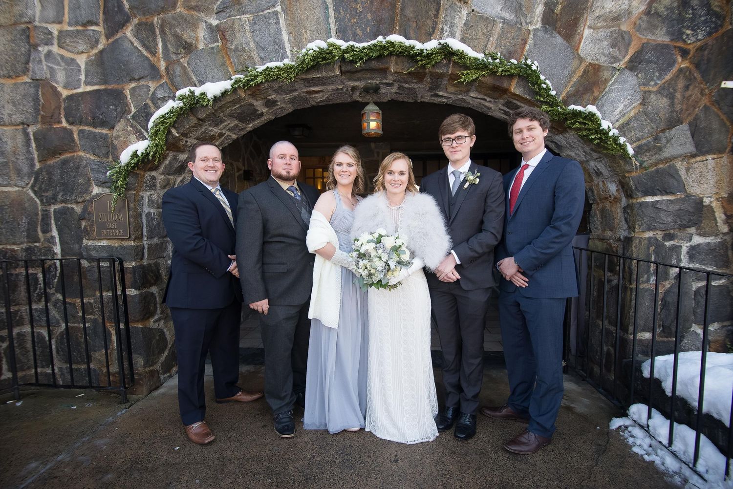 Six people in formal wedding attire smiling before a rustic stone archway with snow.