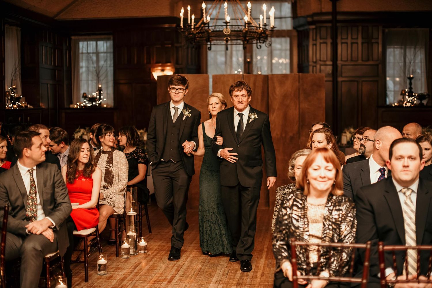 Elegant wedding processional in a wood-paneled ballroom with chandelier lighting, guests seated as bridal party walks the aisle.