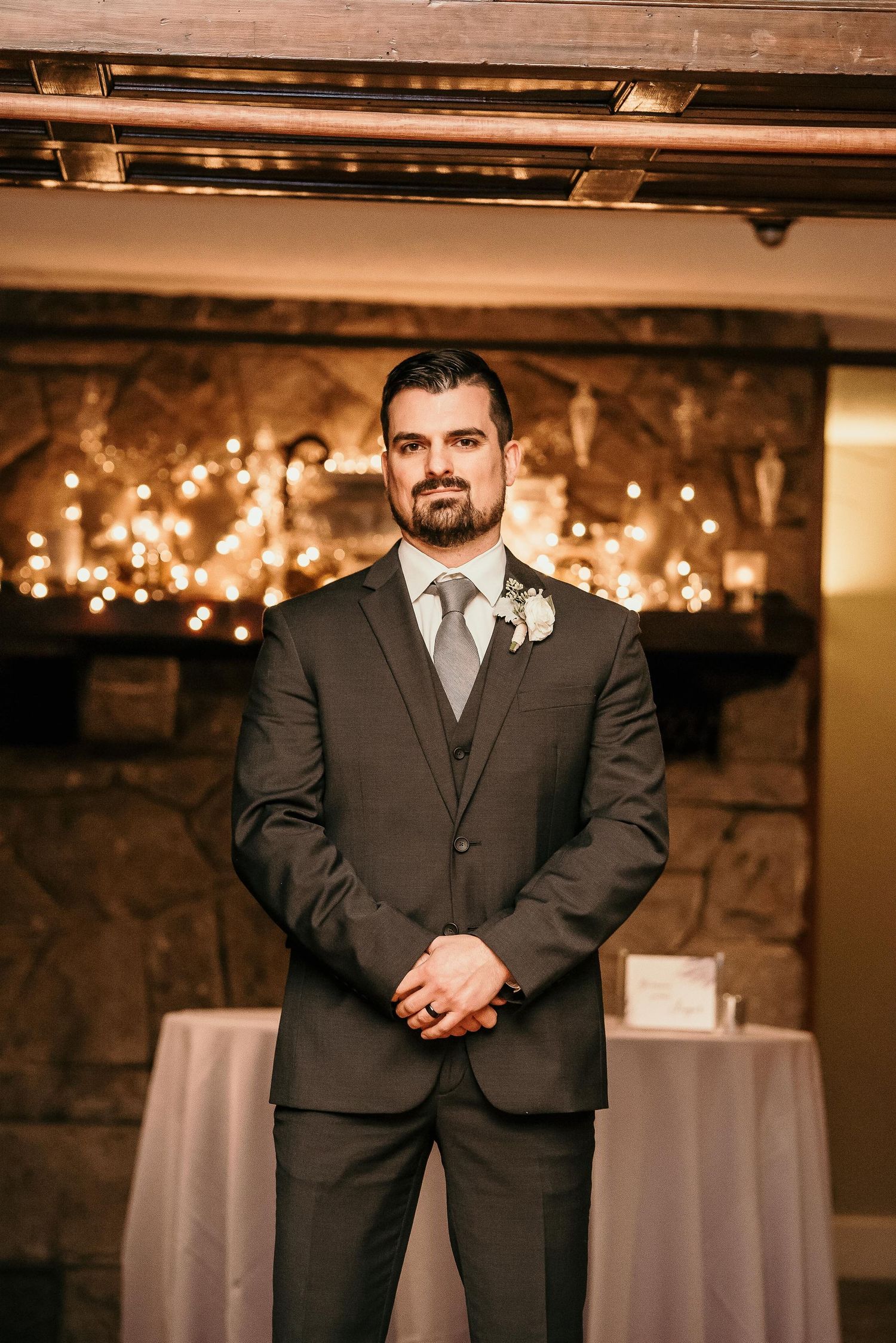 Groom in dark suit with boutonniere stands before stone fireplace with warm string lights at wedding venue.