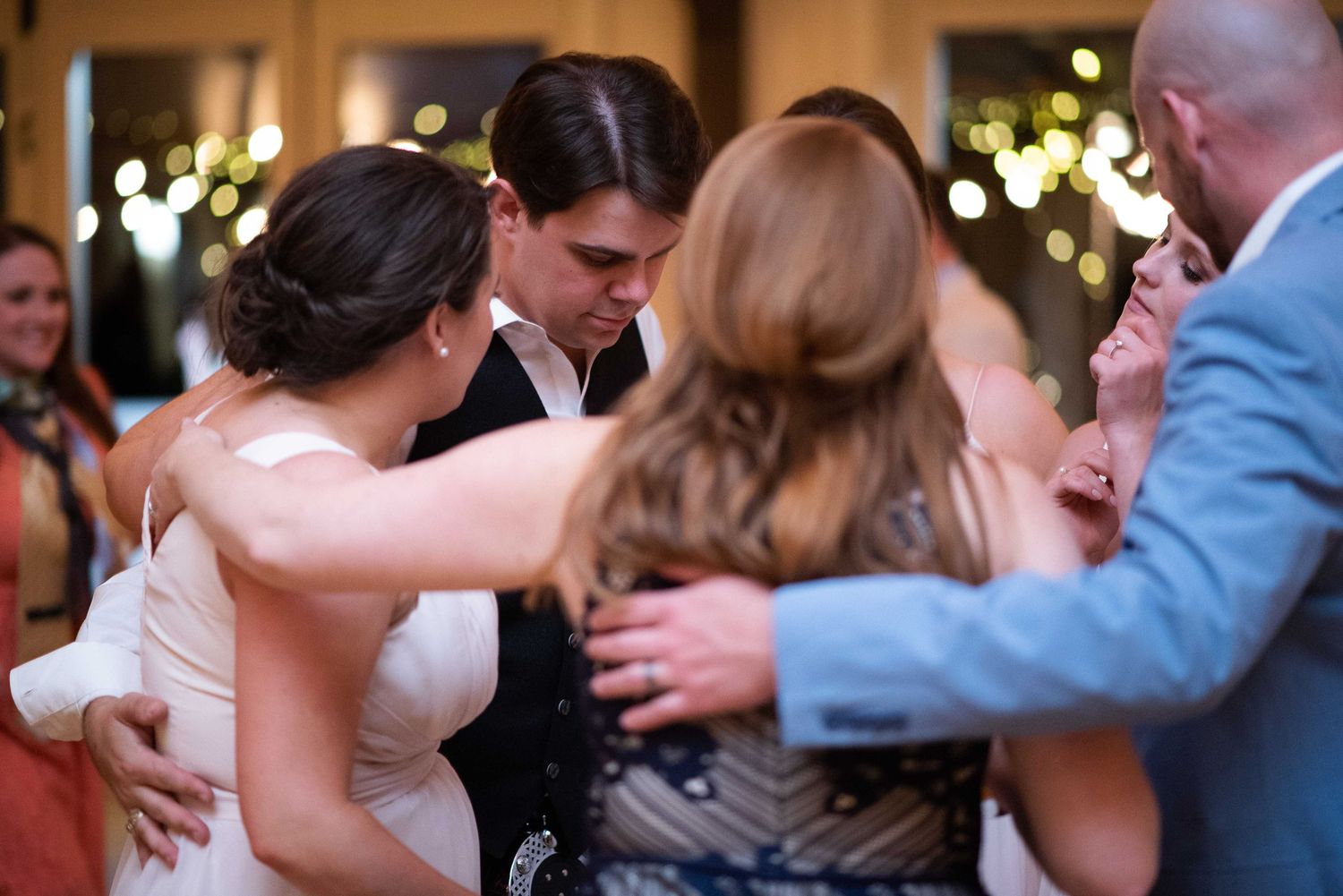Bride and groom share a joyful group hug with wedding guests on the dance floor at a reception.