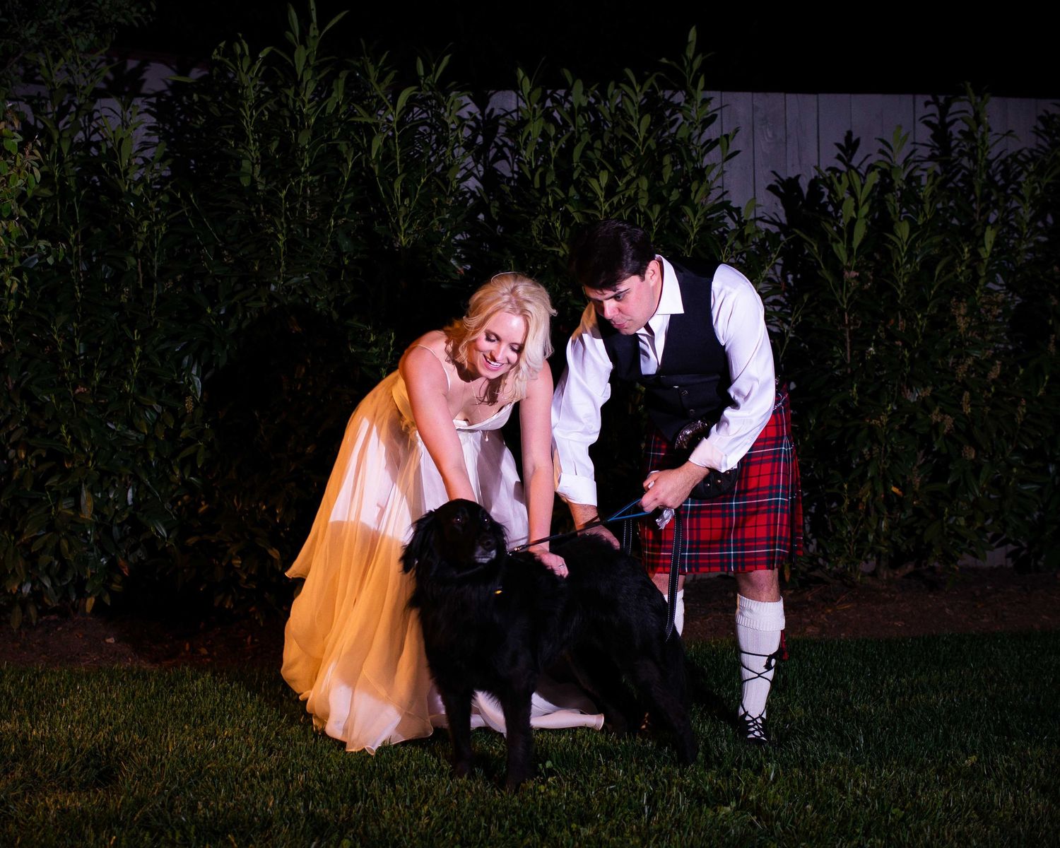 Bride in yellow gown and groom in red tartan kilt petting a black dog outdoors at night.