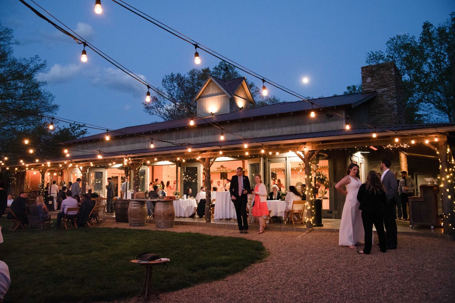 Outdoor wedding reception at dusk with string lights illuminating a rustic barn venue and elegantly dressed guests.