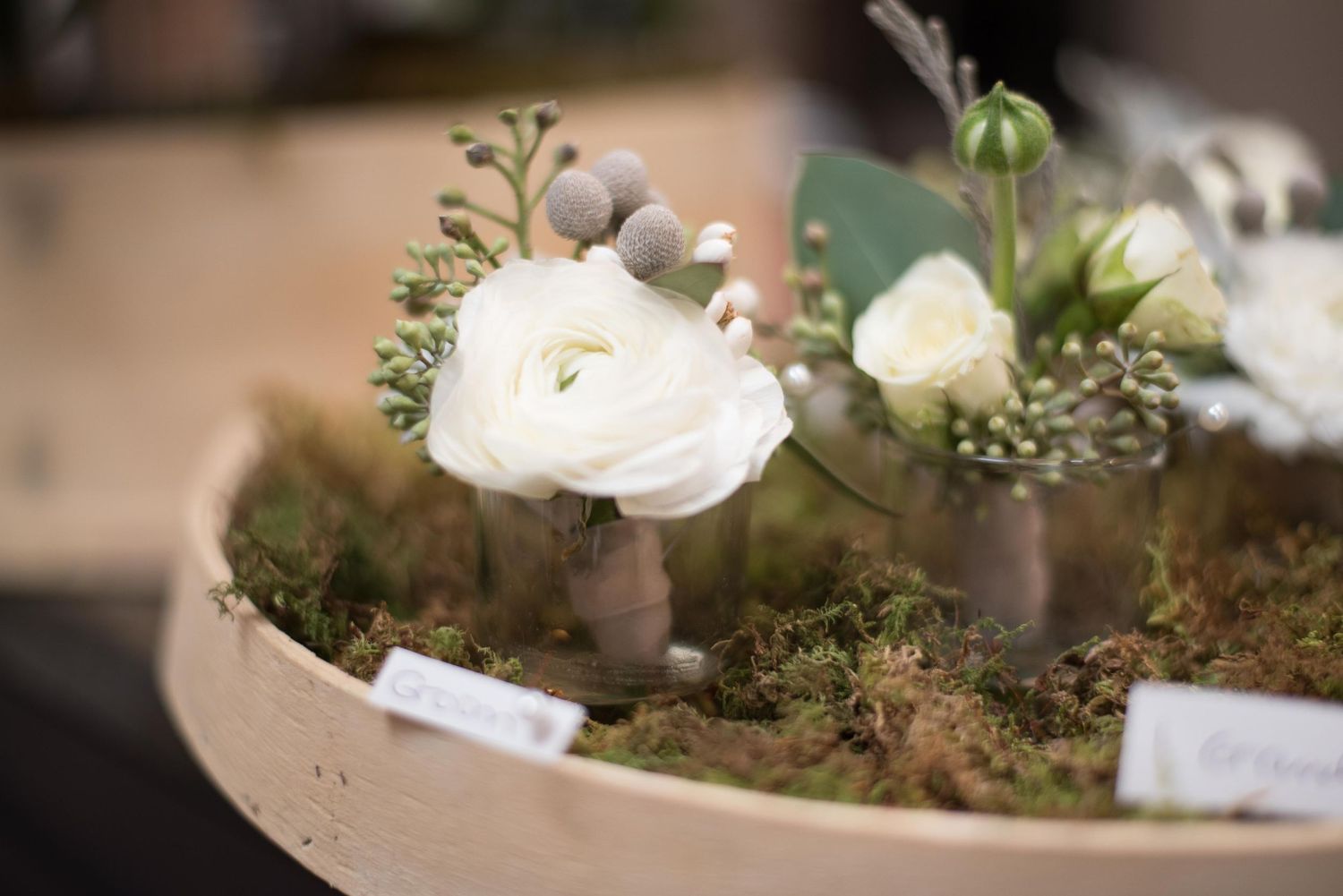 Elegant white ranunculus and roses arranged in small vases with moss in a wooden bowl for a wedding centerpiece.