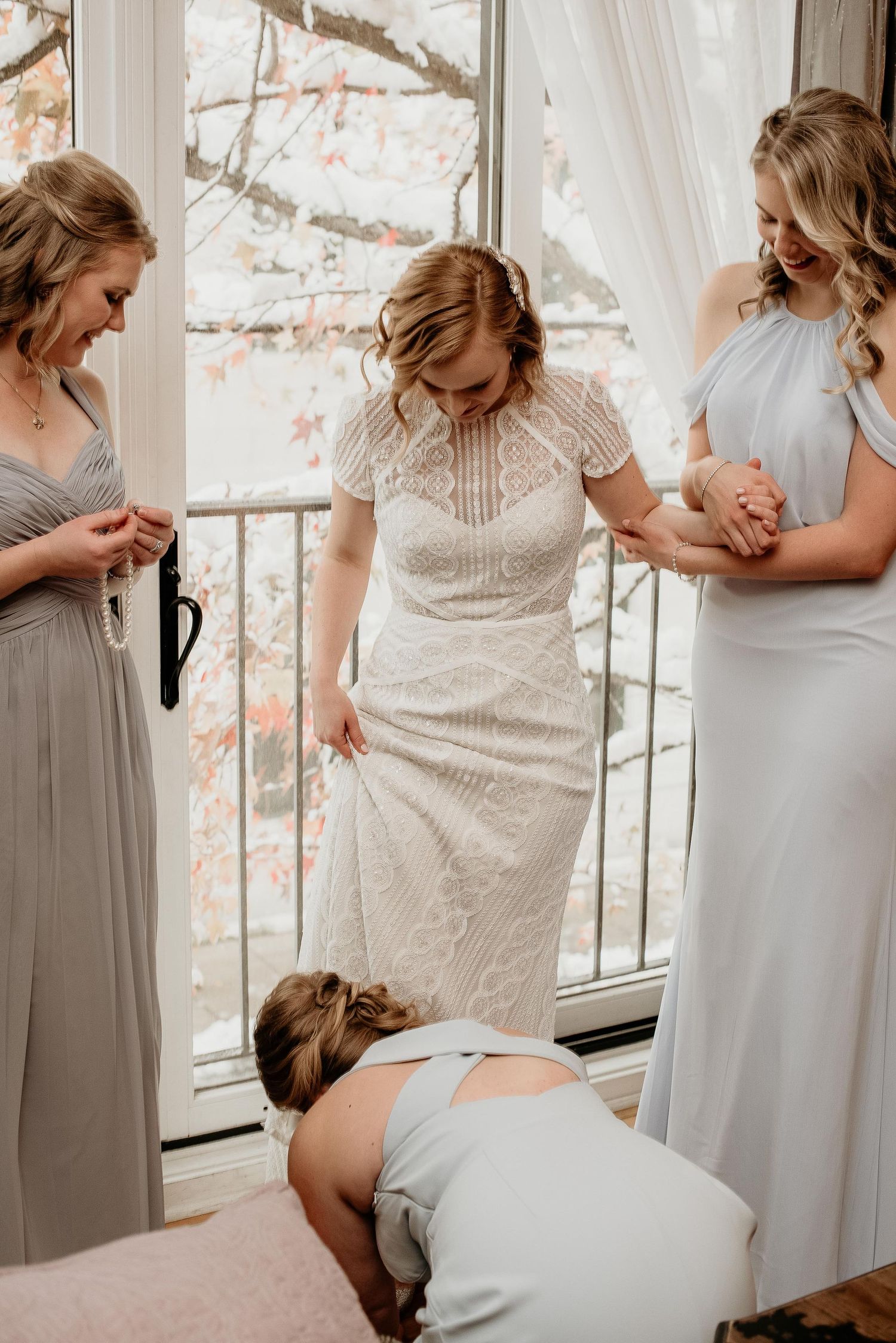 Bridesmaids helping bride into white lace wedding dress near bright window with autumn trees in background.
