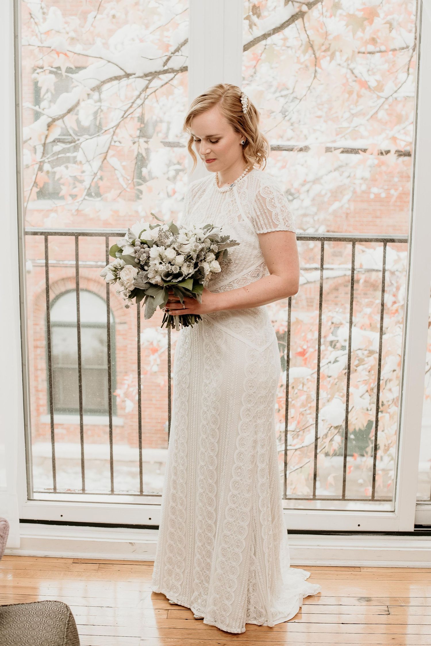 Bride in white lace gown holding bouquet of white flowers and greenery, standing before large windows with snowy trees.