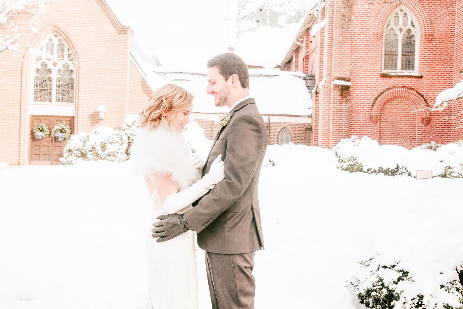 Bride and groom embrace in snow outside a red brick church during a romantic winter wedding ceremony.