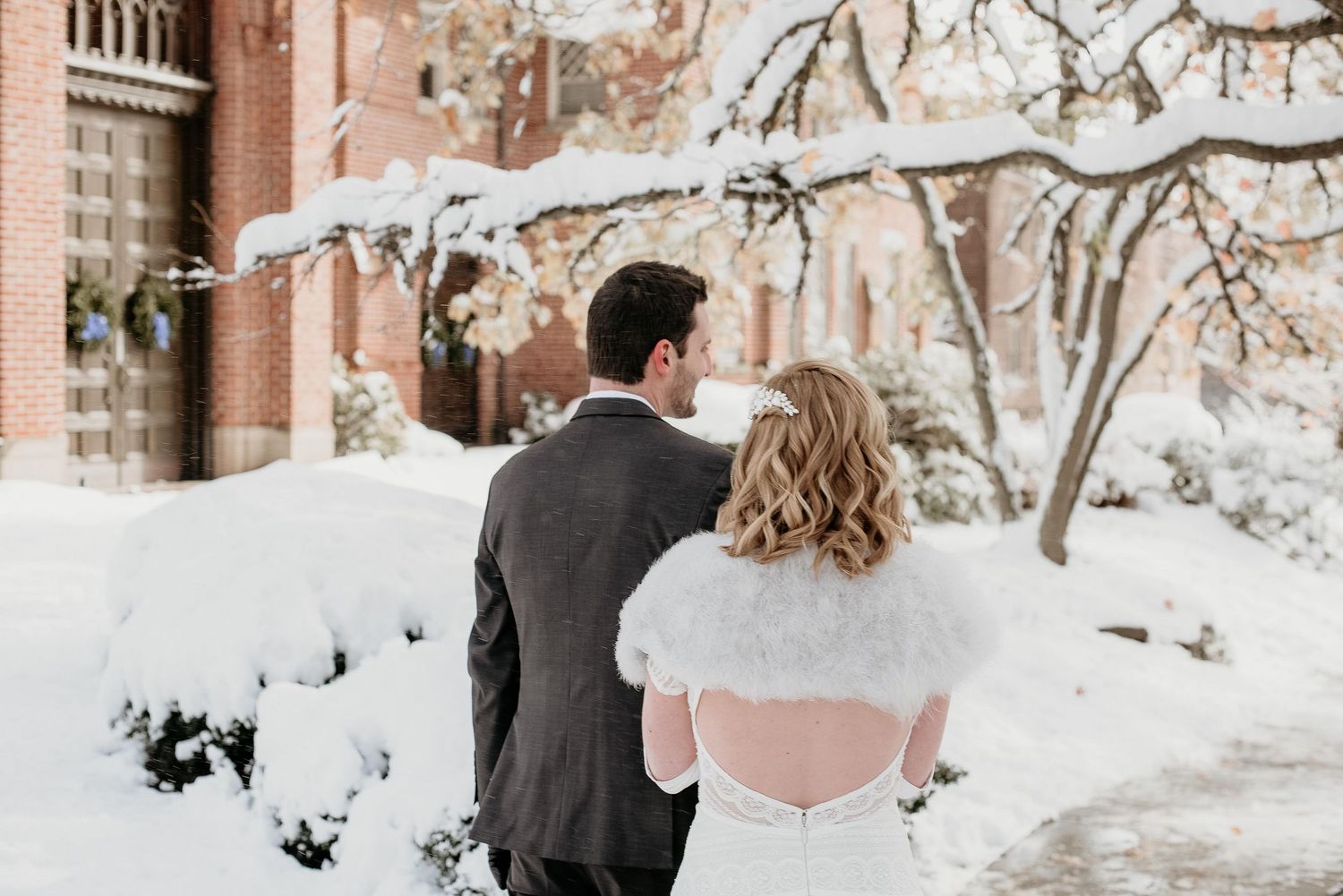 A bride and groom walk together in a snowy winter landscape, the bride wearing a white fur shawl and wedding dress.