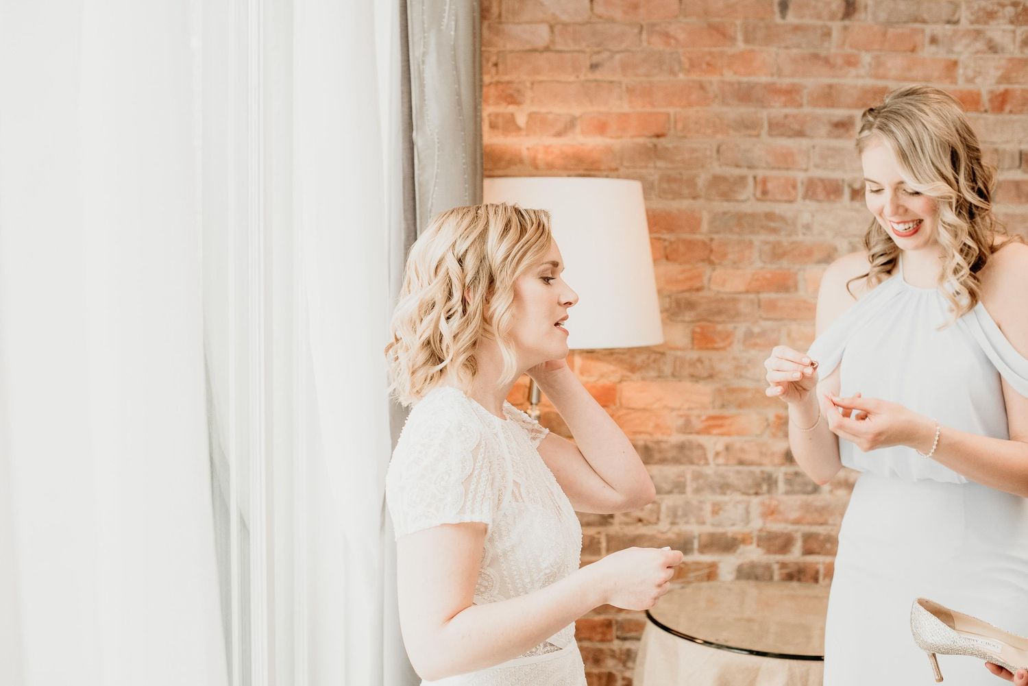 Two blonde women in white dresses laughing and sharing a joyful moment in a room with exposed brick walls.