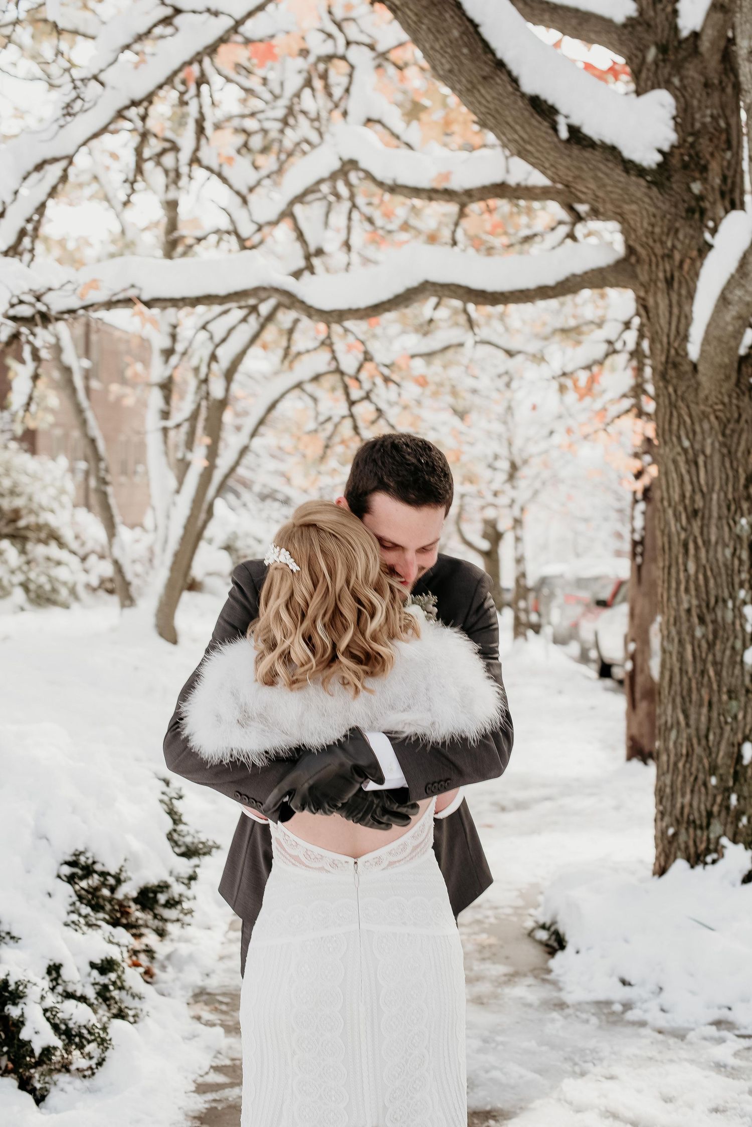 Bride and groom embrace in a snowy winter wonderland, bride wearing a white fur stole and lace gown.