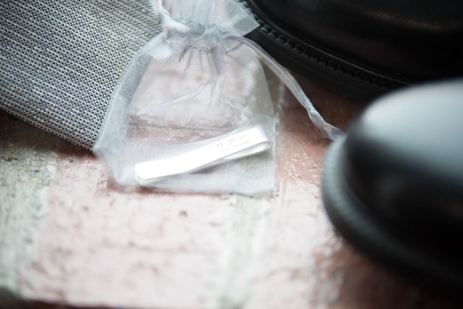 Silver tie clip in a sheer organza gift bag on a marble surface with dress shoes in background.