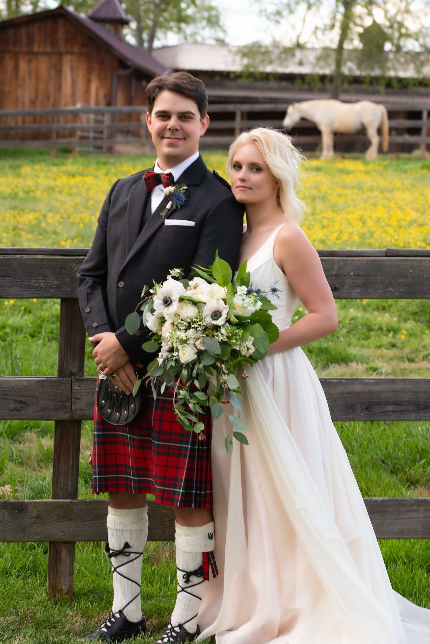 Bride and groom in Scottish kilt pose by wooden fence at farm wedding with white horse grazing in background.