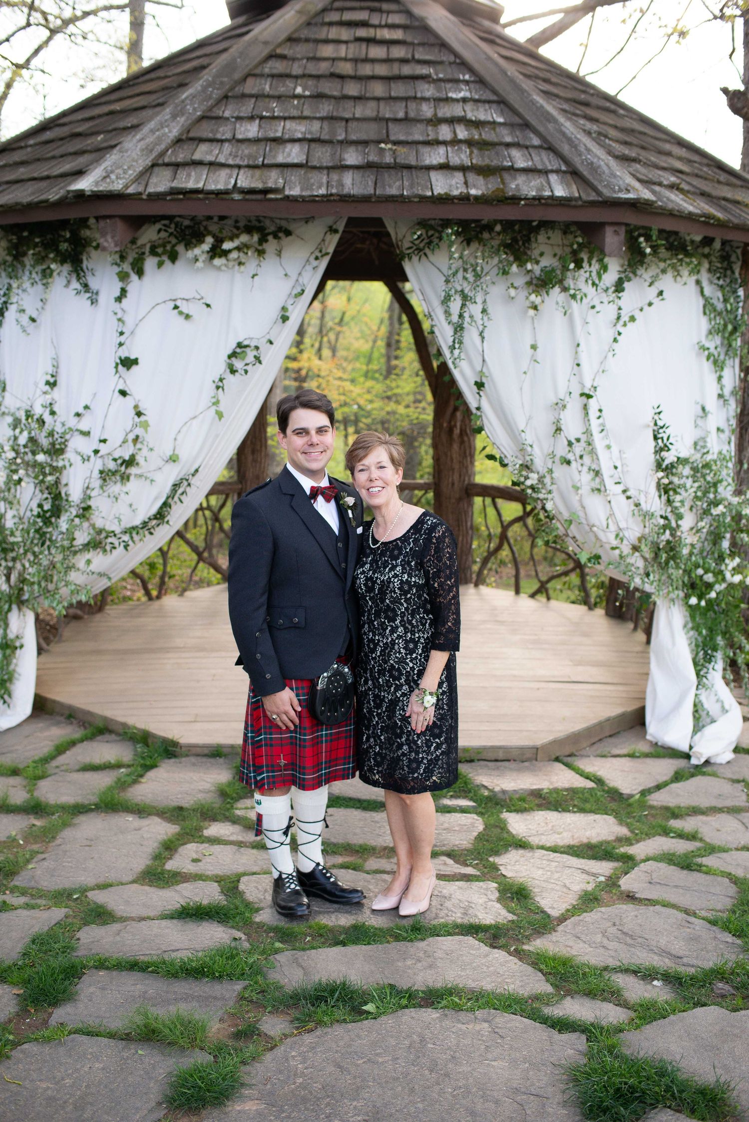 Couple in formal attire, man in kilt, posing before decorated gazebo with white drapes and greenery.