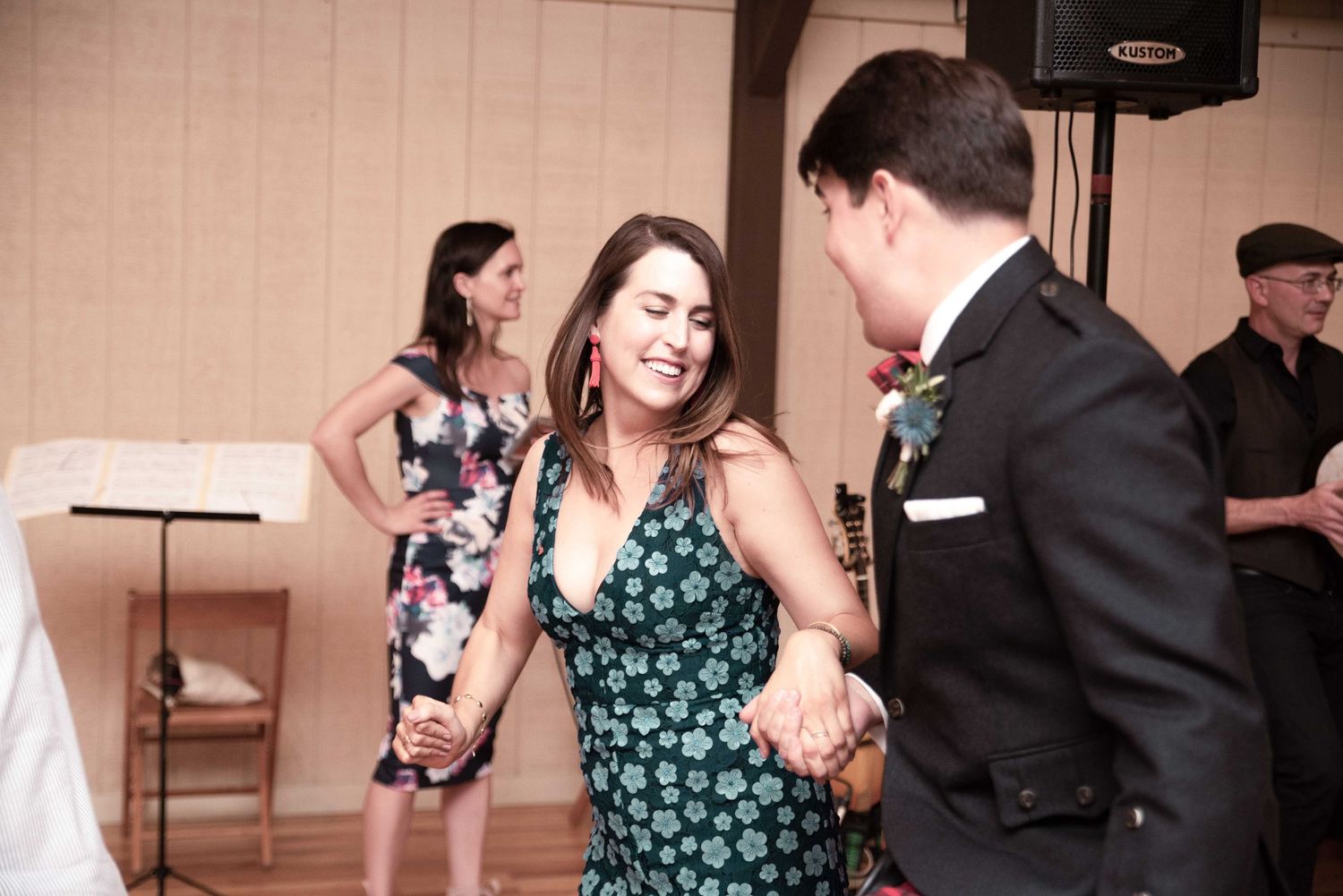 Couple dancing joyfully at a wedding reception, woman in floral dress laughing, guests watching in background.