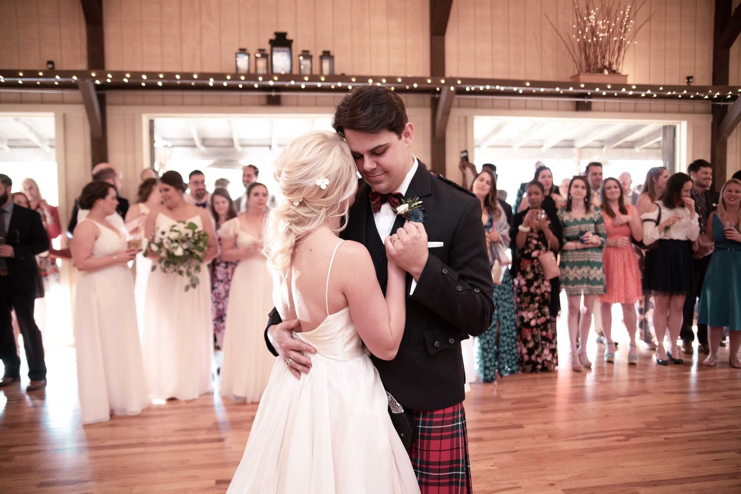 Bride and groom share first dance at rustic barn wedding reception, groom wearing traditional Scottish tartan kilt.