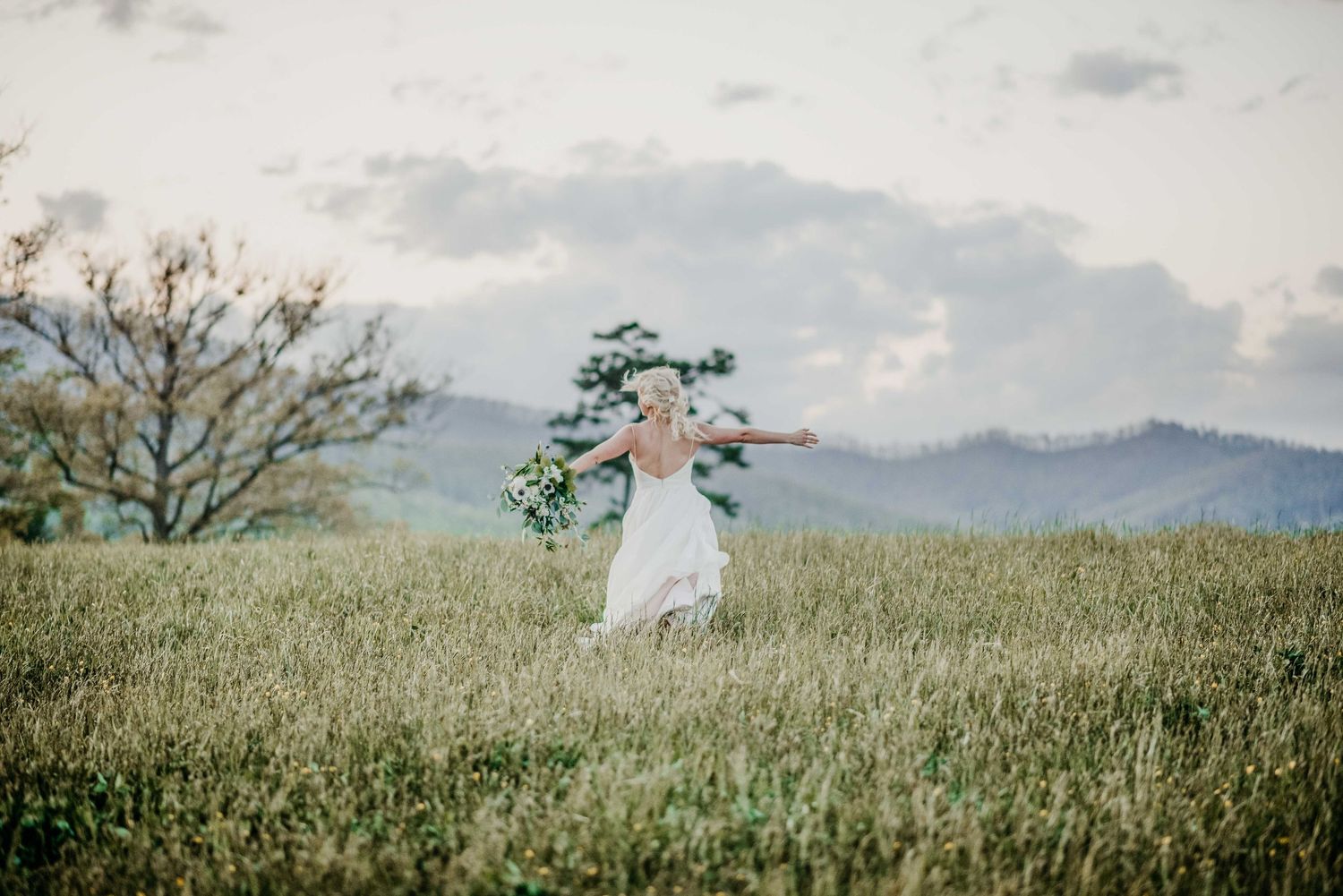 Bride in white dress running through green meadow with bouquet, mountains and cloudy sky in background.