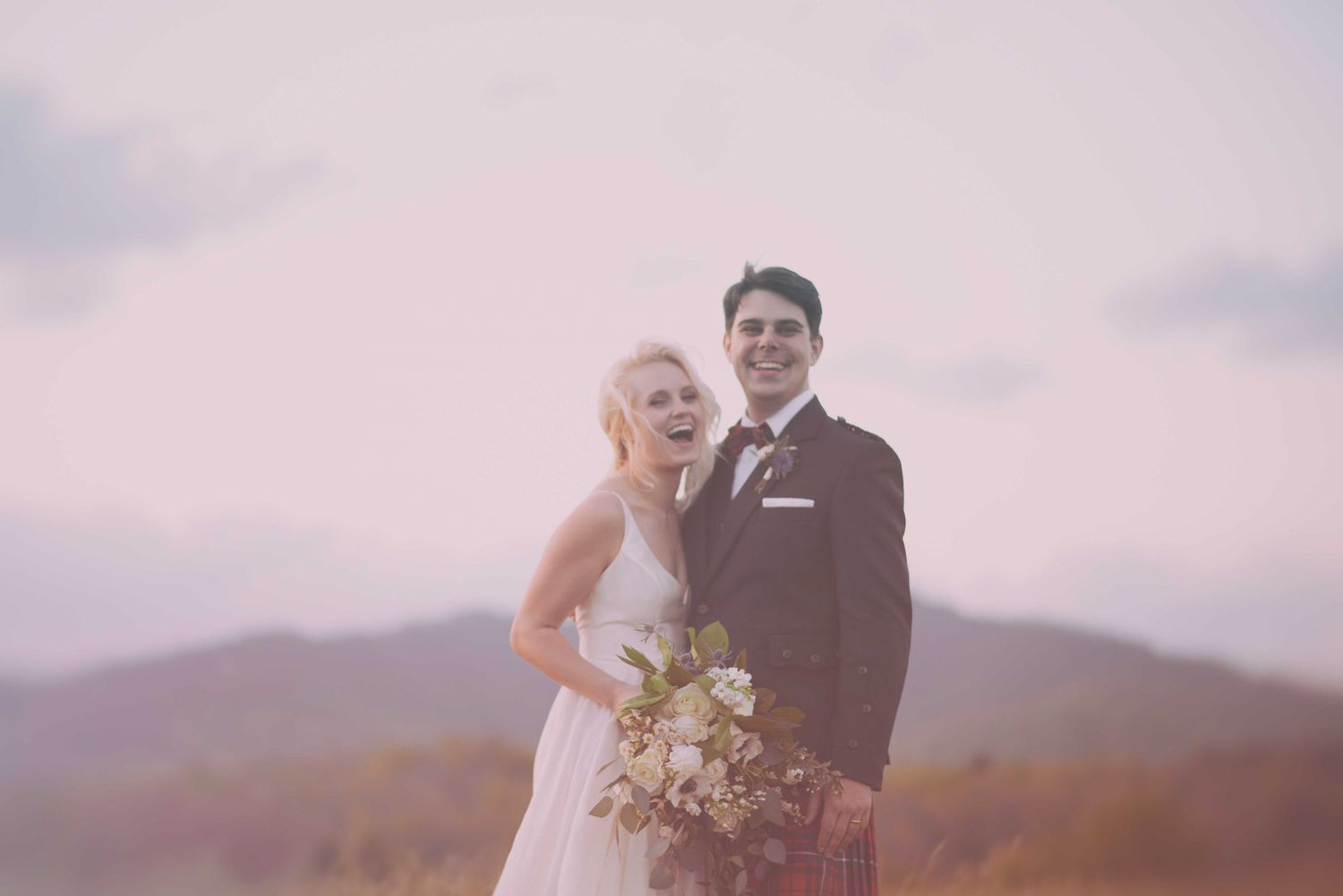Happy bride and groom posing outdoors with mountain backdrop, bride holding bouquet in white gown, groom in dark suit.